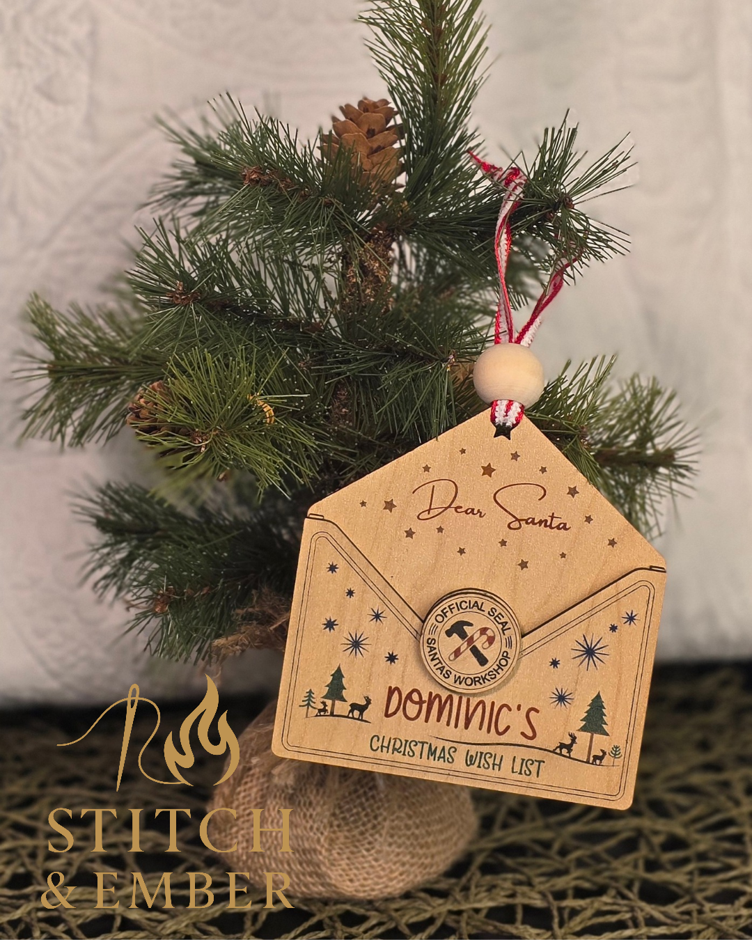 A small Christmas tree with a wooden tag hanging from a red and white string. The tag reads "Dear Santa," and has a printed seal that says "Official Seal Santa's Workshop." The tree is decorated with a pine cone and a burlap-like base. The background