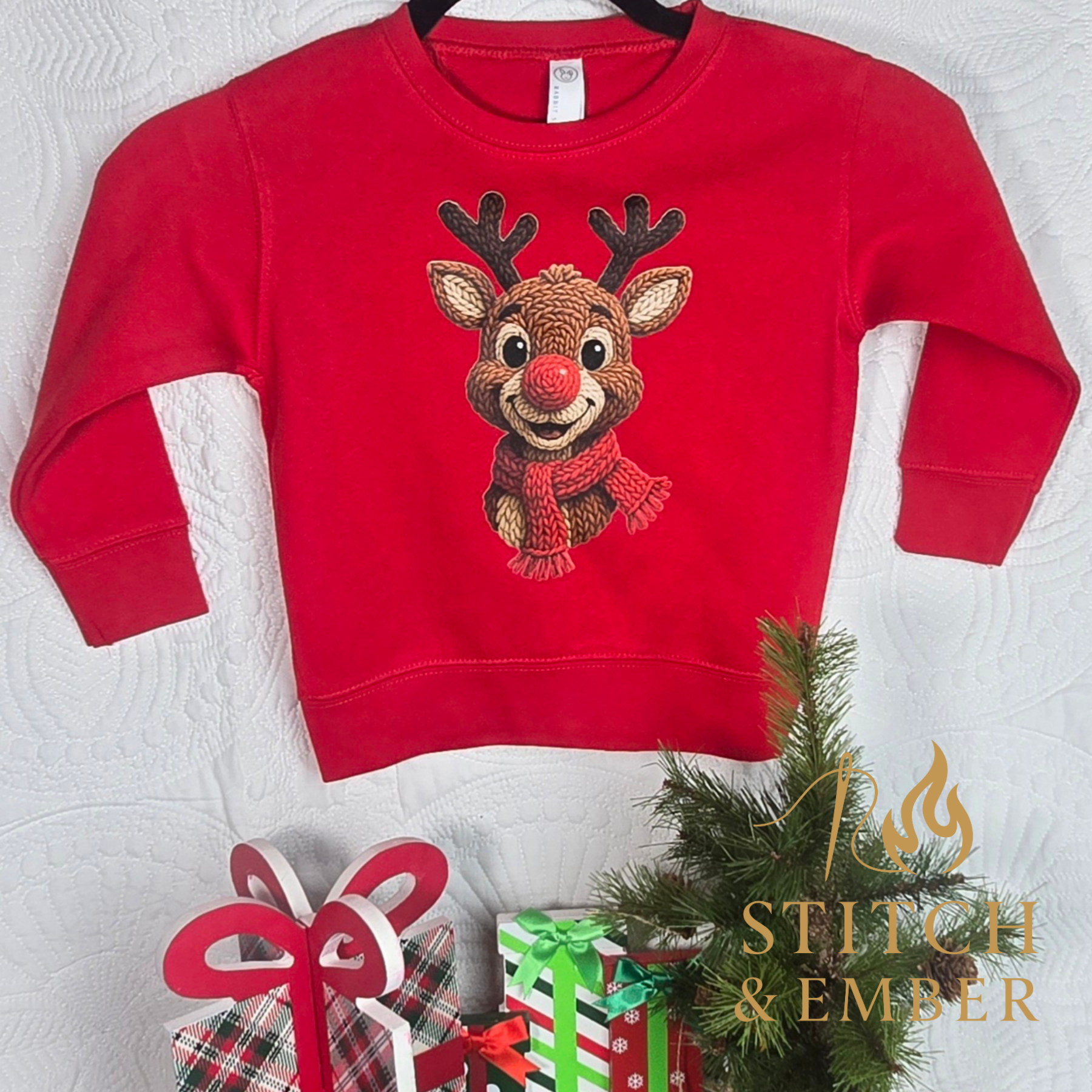 Red Christmas sweatshirt with a reindeer wearing a red nose and scarf, displayed on a white quilted background, accompanied by small gift boxes, a small pine branch, and a logo reading 'Stitch & Ember'.