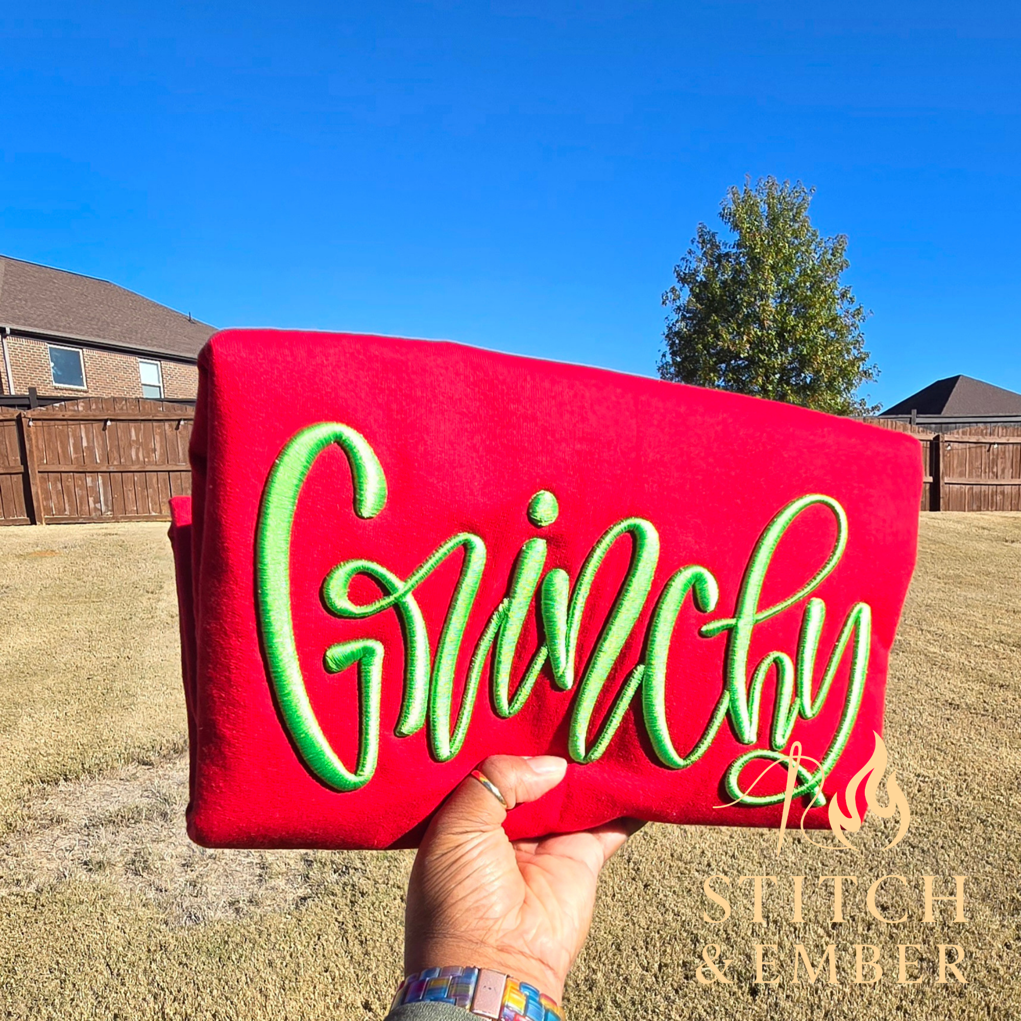 Hand holding a red clutch with green neon 'GlamChic' embroidery, in a backyard with a fence and trees under a blue sky.