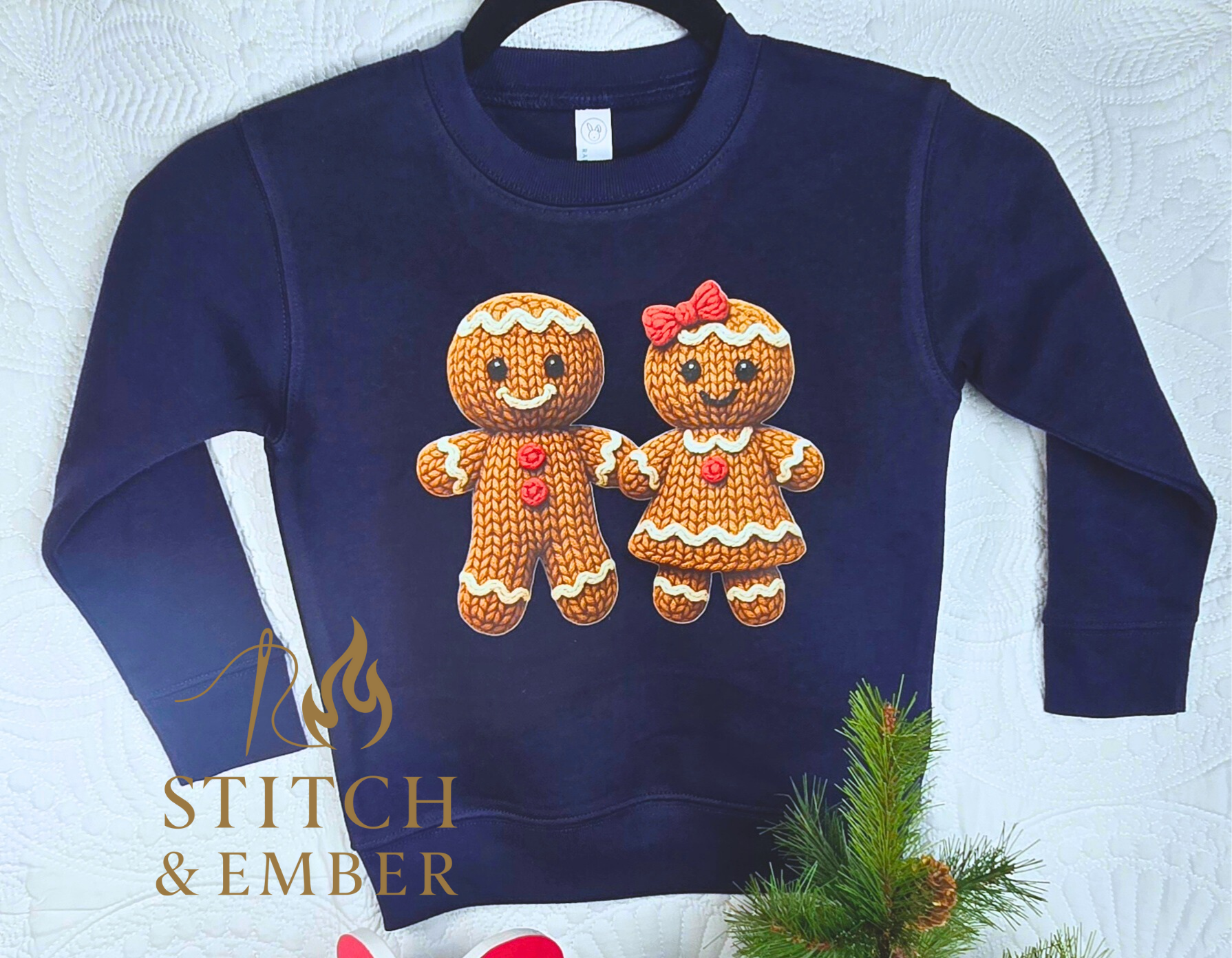 A navy blue sweatshirt with adorable embroidered gingerbread cookies, a boy and a girl, holding hands and decorated with white icing and red buttons, hanging on a black hanger against a white textured background with a small branch of pine with cones