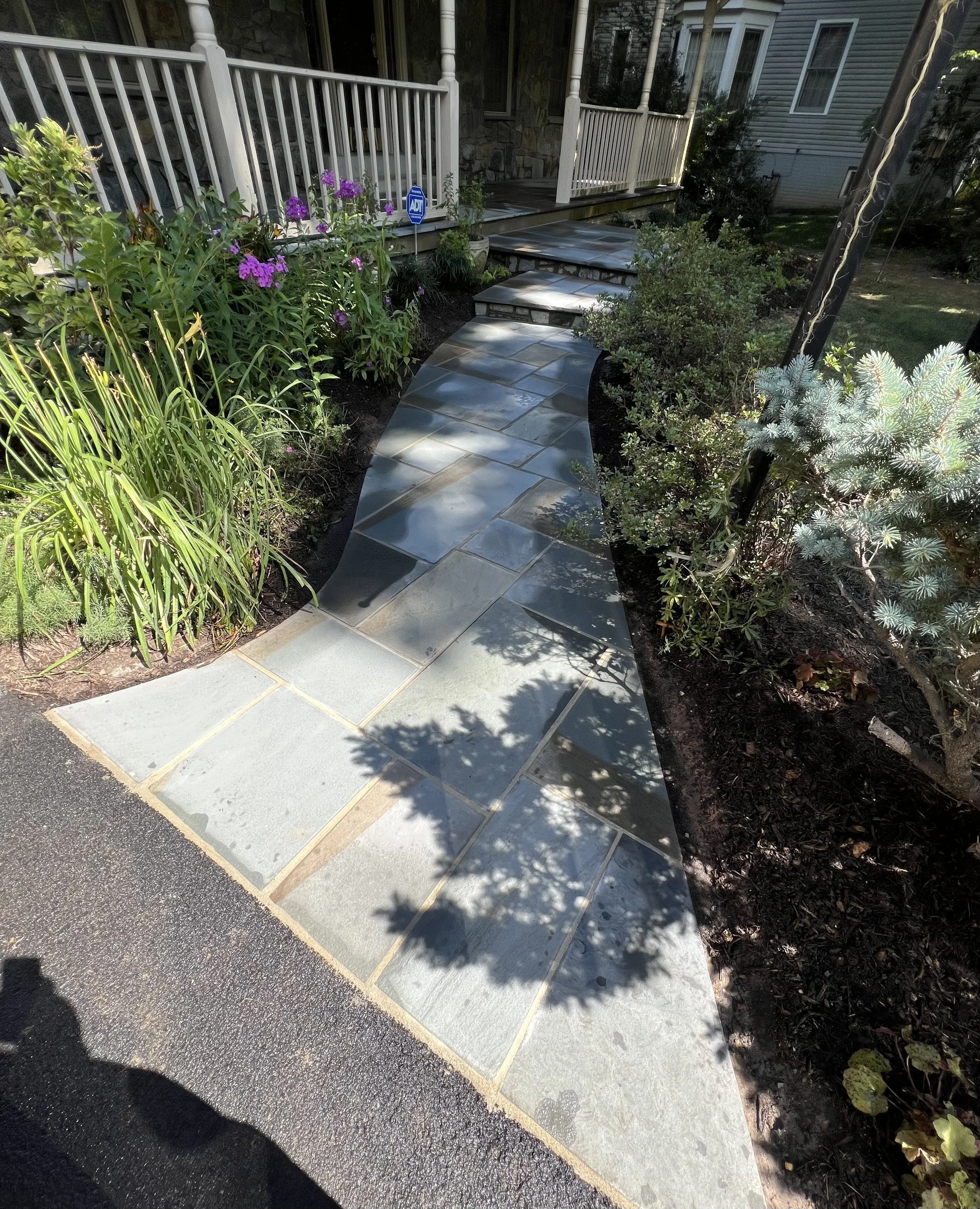 A curved stone pathway leading to a porch surrounded by greenery and flowers.