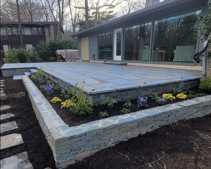 A modern outdoor patio with stone flooring and a raised garden bed, featuring colorful flowers and neatly arranged stones, adjacent to a house with large glass windows.