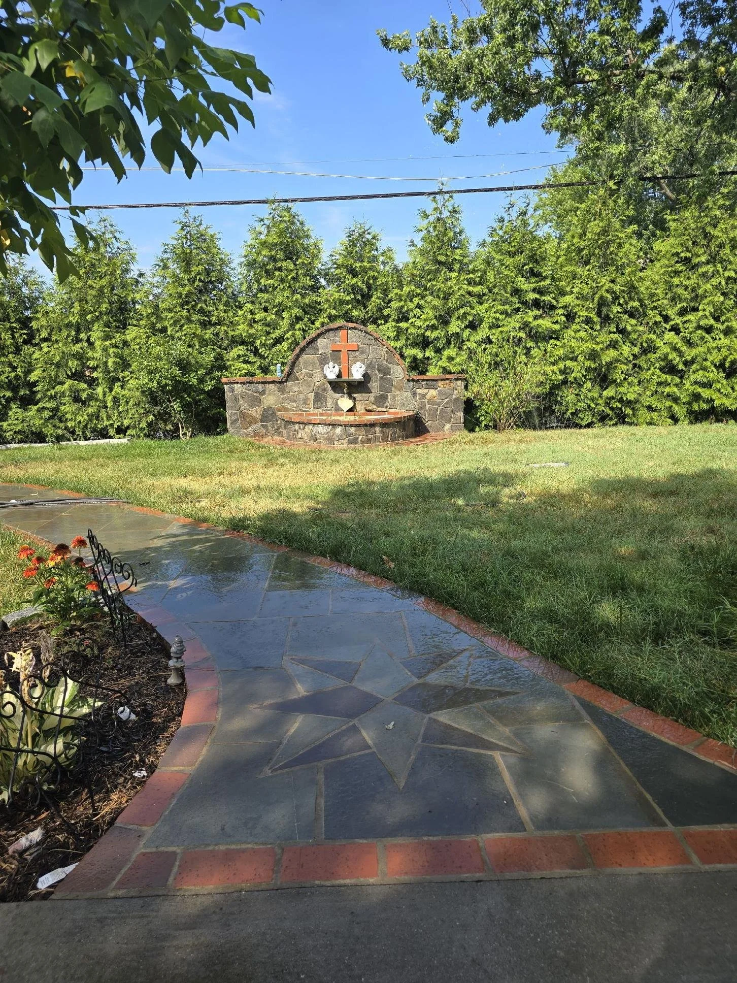 A garden with a stone pathway leading to a stone altar featuring a red cross and two white vases, surrounded by greenery and trees.