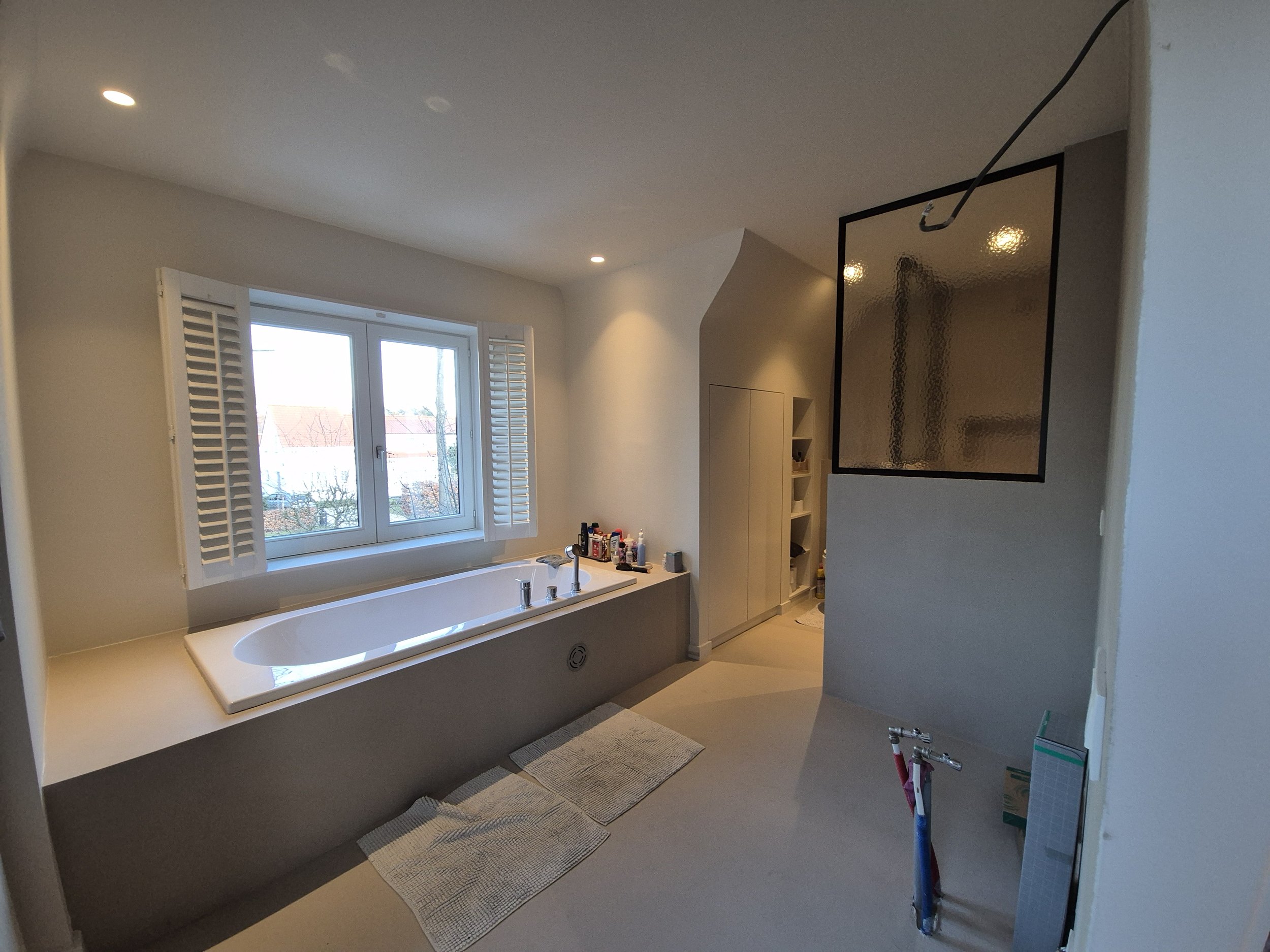 Moderne badkamer met vrijstaand bad naast raam met houten luiken, glazen scheidingswand, handdoeken op de vloer en eenvoudige decoratie.