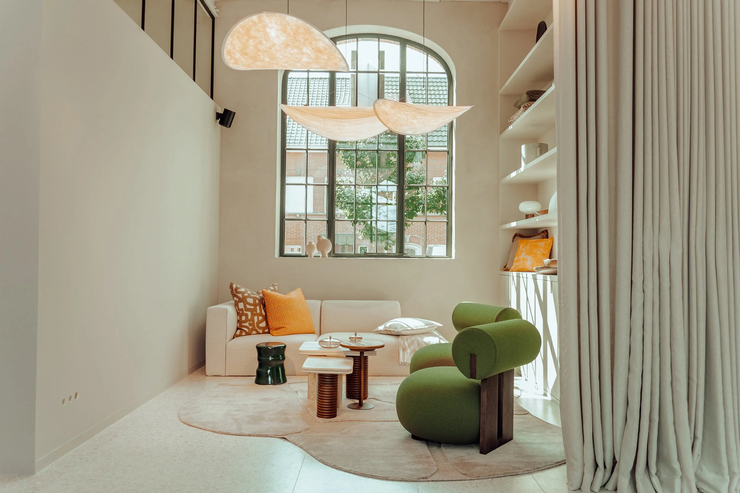 Moderne woonkamer met beige bank, groene fauteuil, decoratieve kussens, ronde tafels, grote boogvormige ramen, en decoratief verlichtingsontwerp aan het plafond.