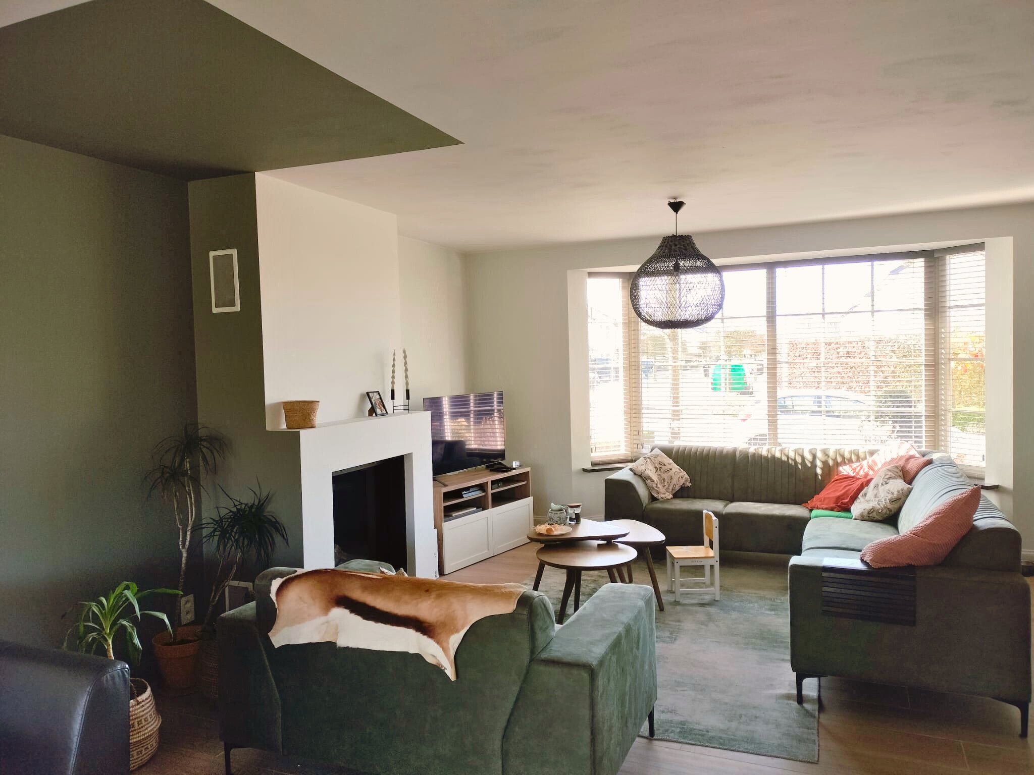 Een woonkamer met groene muren, een zithoek met groene banken en verschillende kussens. Er zijn kamerplanten, een hanglamp, een televisie op een kastje, en een houten salontafel.
