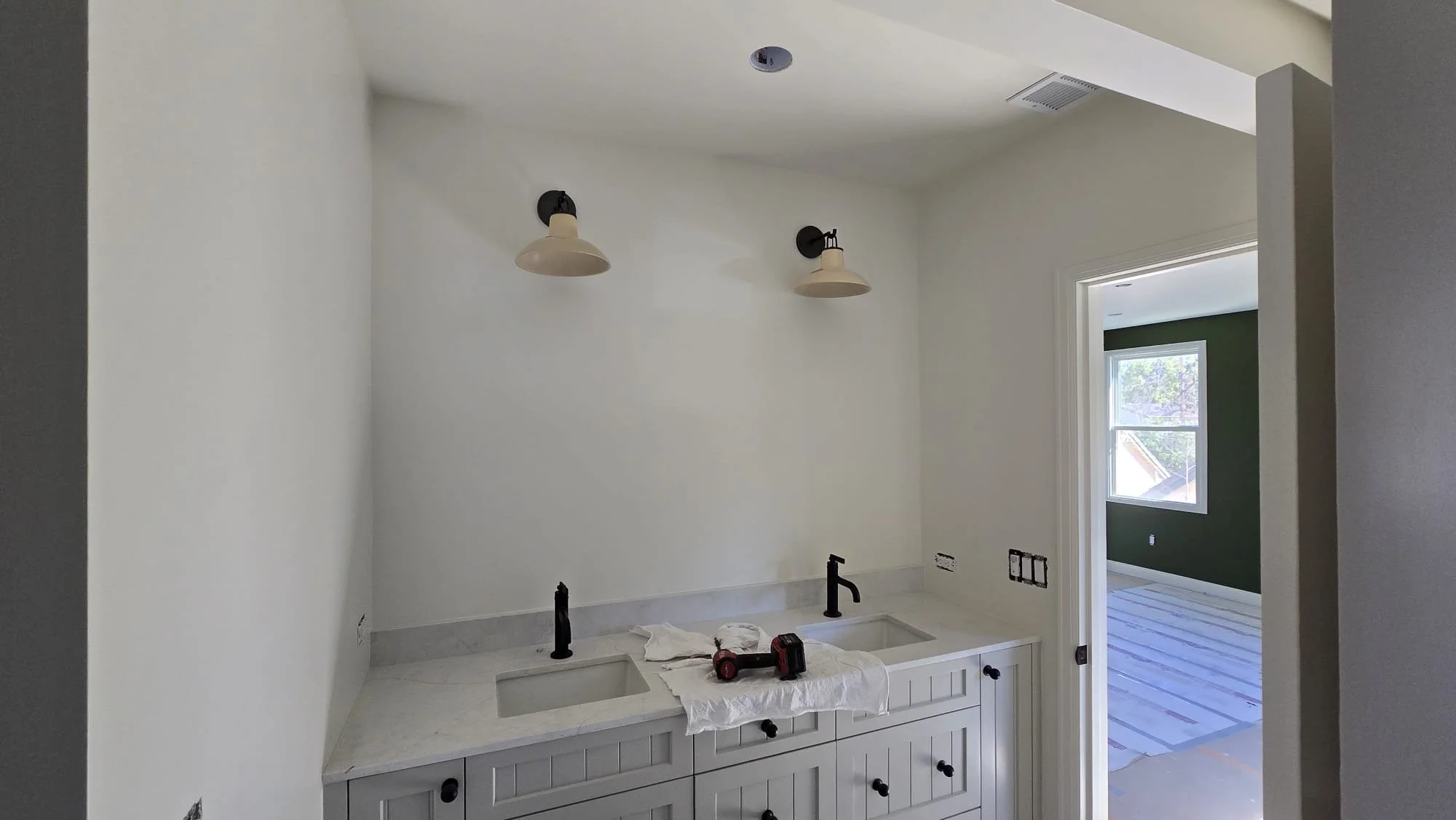 Partially finished kitchen with white cabinets, a marble countertop with a double sink, black faucets, and unfinished wall outlets. There are two wall sconces above the countertop, an opening to a room with green walls and a window. Construction tool