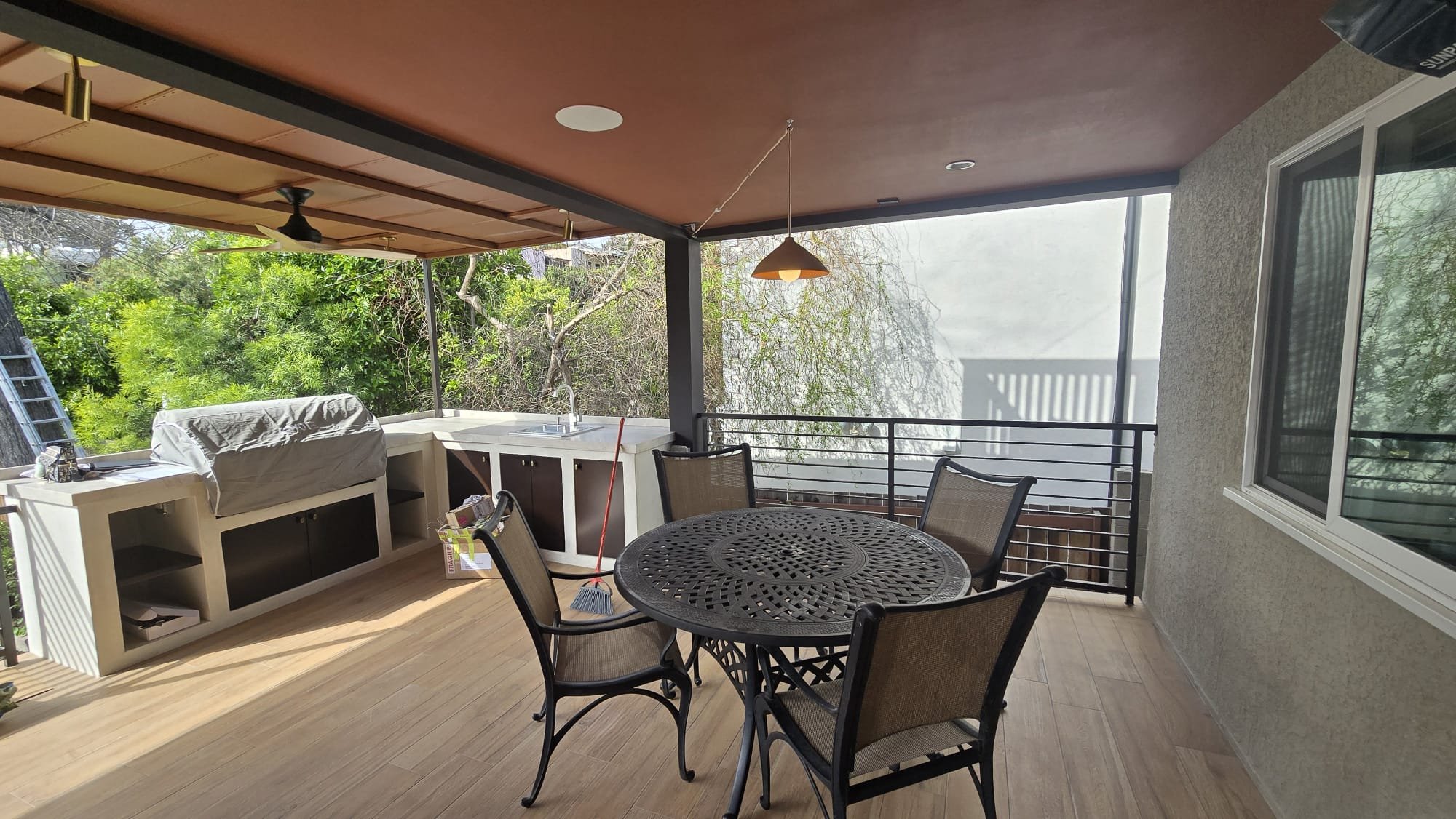 Covered balcony with a round black metal table, four chairs, a small sink, and green trees in the background.