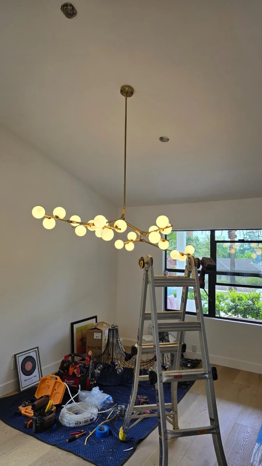 A room under renovation with a modern chandelier hanging from the ceiling, a ladder, tools, and painting supplies on the floor, and a window showing trees outside.
