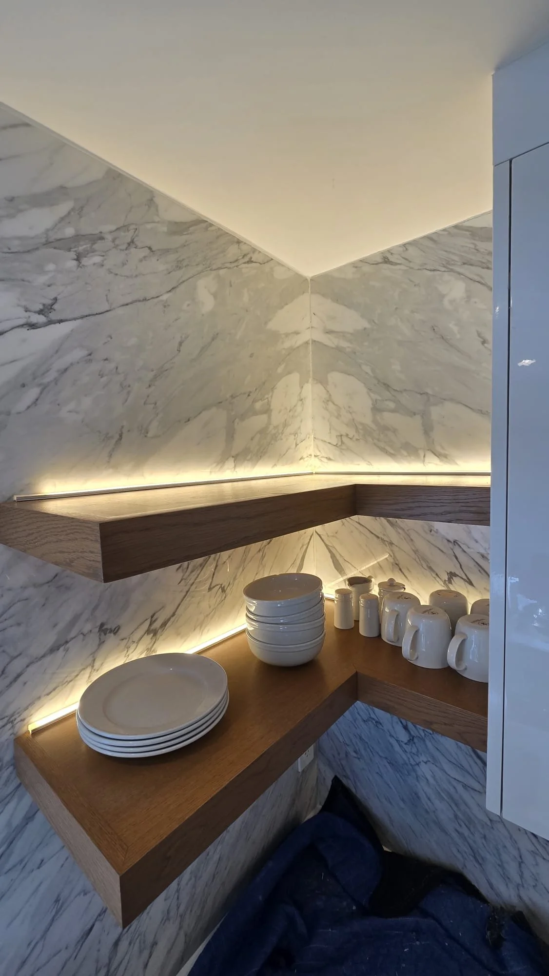 A corner of a kitchen with marble walls and a wooden countertop holding stacked white bowls and cups, and white cups and mugs.