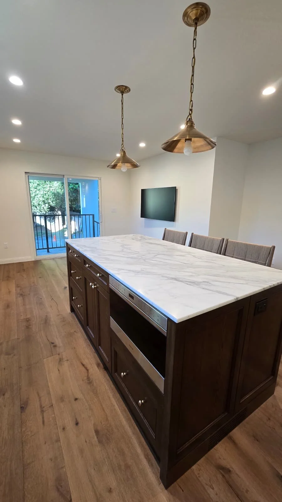Interior of a modern home dining area with a kitchen island featuring a marble countertop, four upholstered chairs, a wall-mounted flat-screen TV, sliding glass door leading to a balcony, and two brass pendant lights hanging from the ceiling.