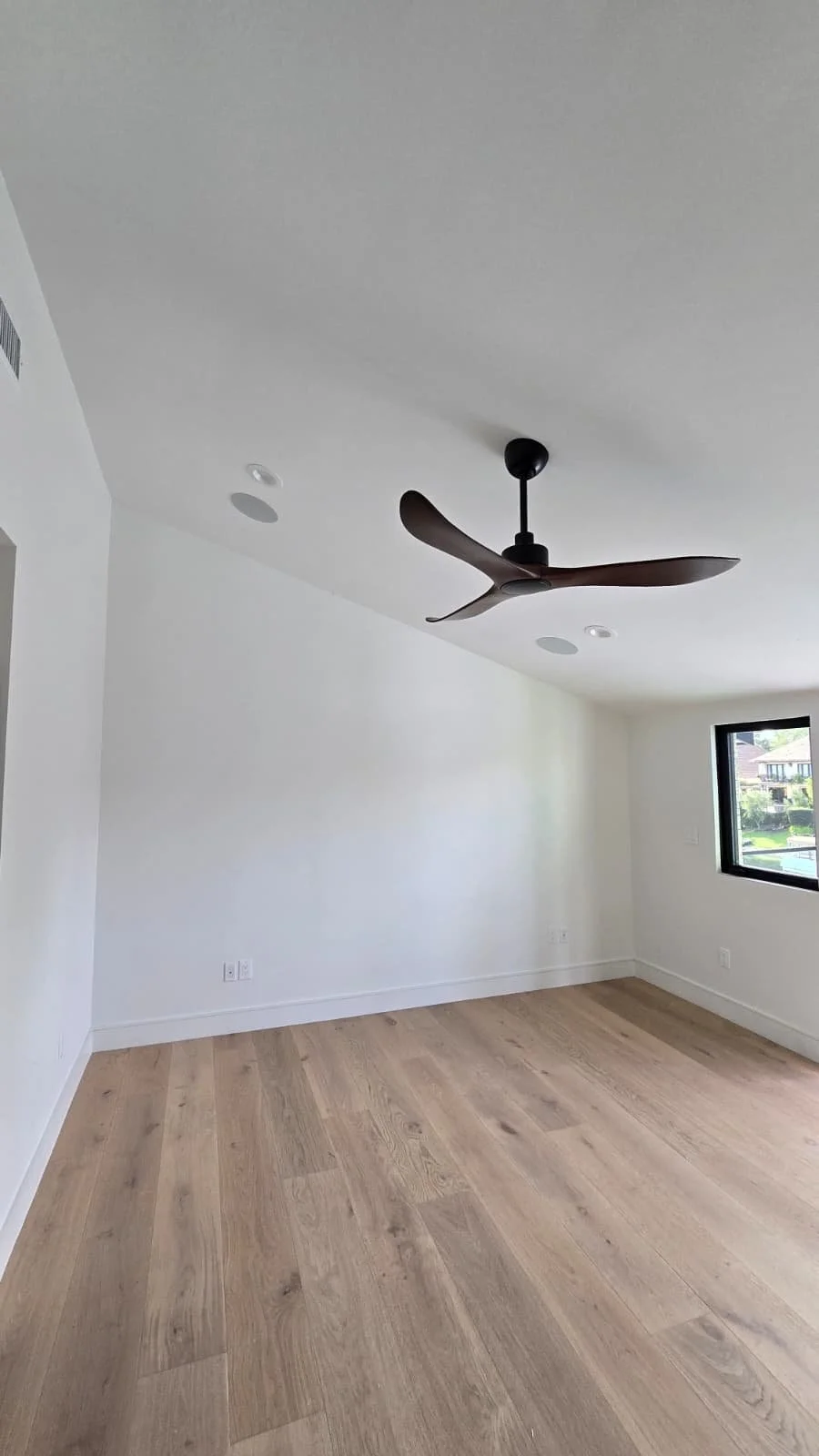 Empty room with white walls, wooden floor, black ceiling fan, and a window showing neighboring houses.