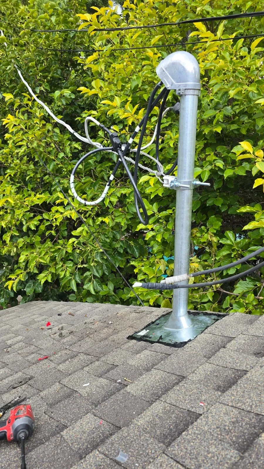 A roof with a metal chimney pipe secured to a roof mount, surrounded by trees and bushes, with electrical wires attached to the pipe and a power drill on the roof.