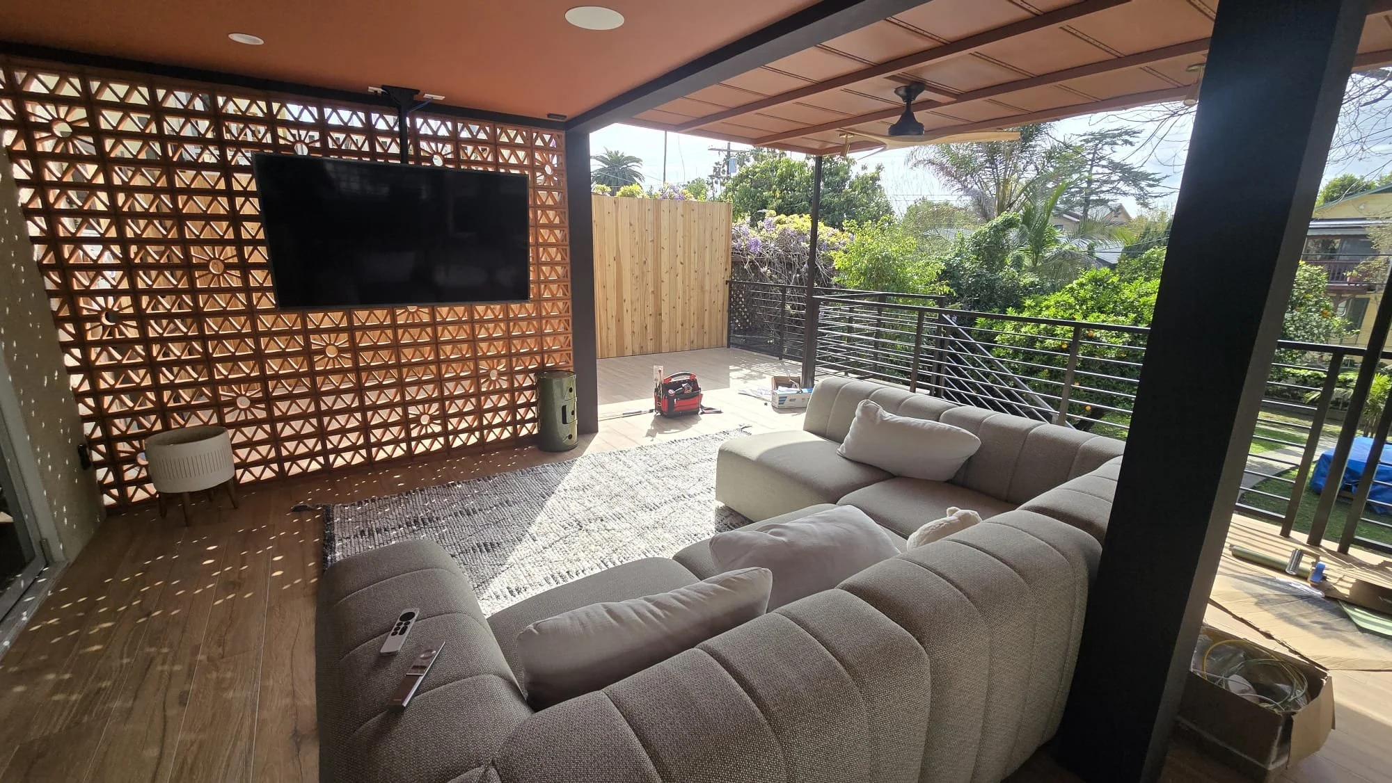 Outdoor balcony with a large sectional sofa, a wall-mounted TV, and view of greenery and trees outside.