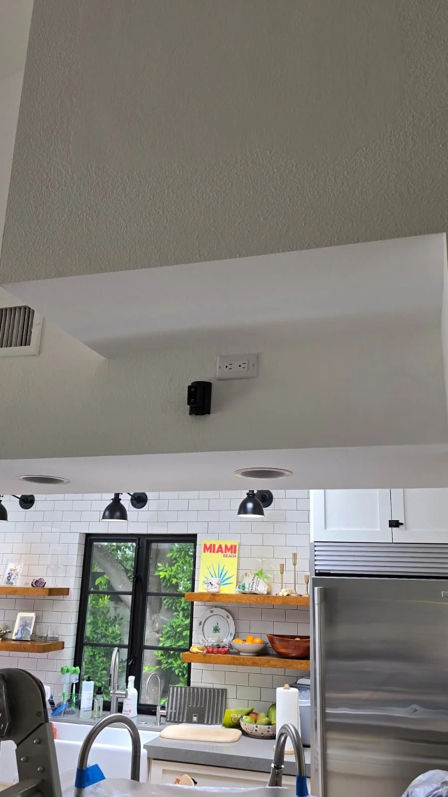 Kitchen with white subway tile backsplash, open wooden shelves, a window with green foliage outside, and various kitchen items including a fruit bowl, soap bottles, and a kitchen towel.