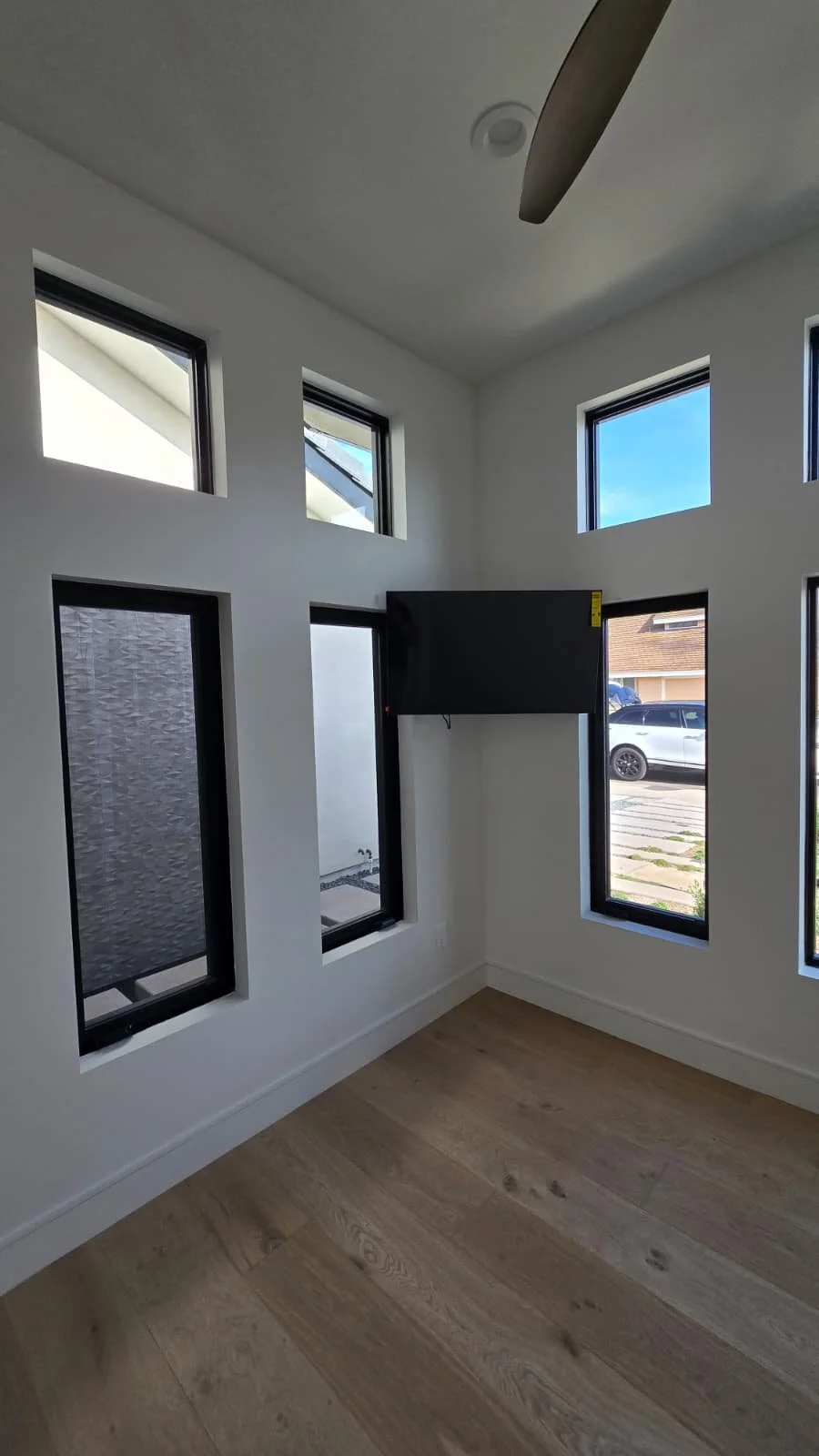 Empty room with multiple small rectangular windows, a wall-mounted TV, light hardwood flooring, and white walls.
