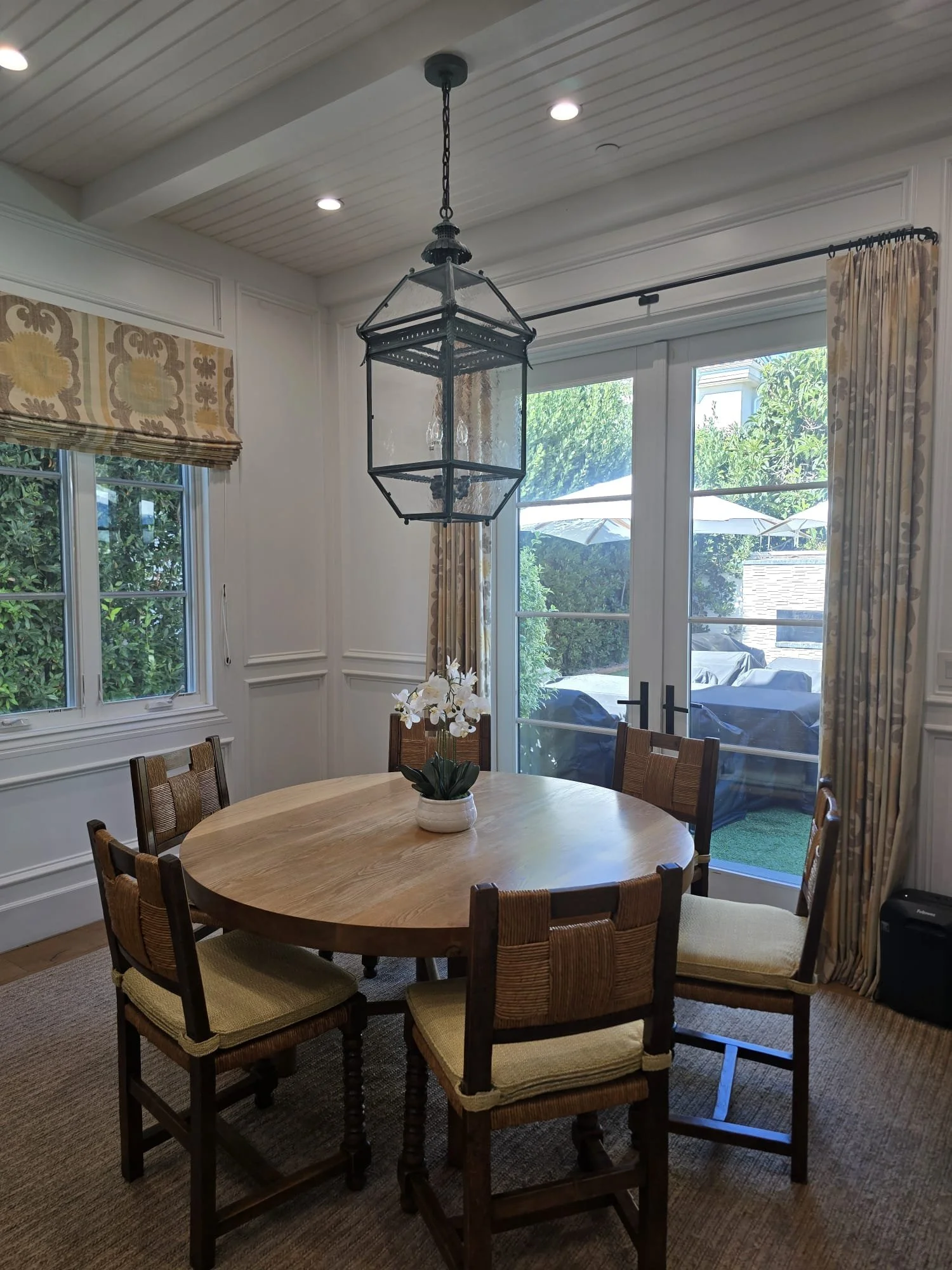 A dining room with a round wooden table surrounded by six wood and woven-straw chairs. There is a white potted orchid in the center of the table. Large windows with patterned curtains allow natural light. A decorative lantern-style light fixture hang