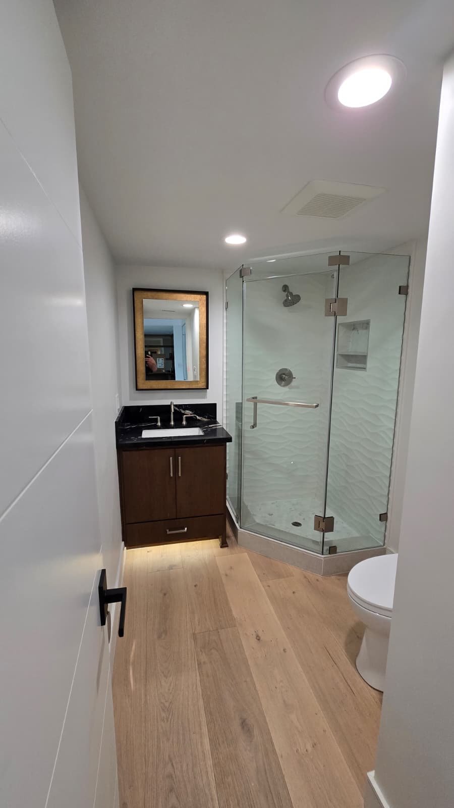 A small modern bathroom with wood floor, white walls, a glass shower stall, a wooden vanity with black countertop, a mirror with a wooden frame, and a white toilet.