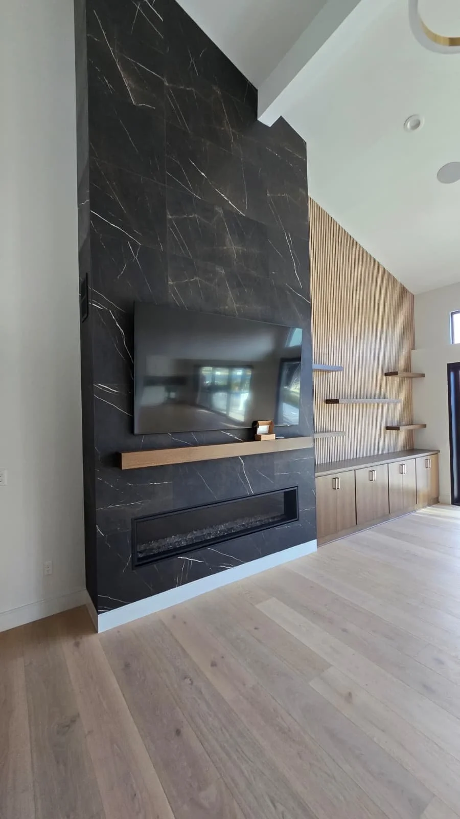Modern living room with a black marble fireplace wall, a mounted flat-screen TV, a wooden shelf below the TV, a built-in electric fireplace, and a wall with vertical wooden slats with floating shelves.