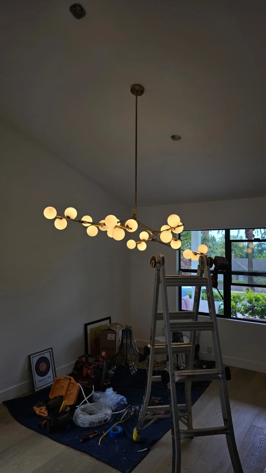 Modern chandelier with multiple glowing globes hanging from the ceiling in a room under renovation, with tools, ladder, and art supplies on the floor and a window in the background.
