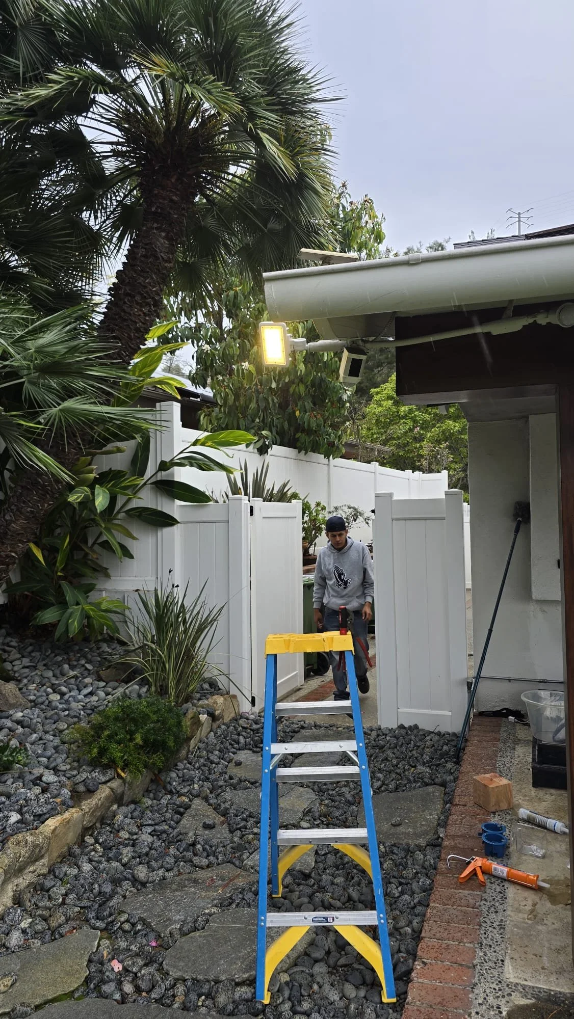 A person standing on a step ladder in a backyard, surrounded by plants and trees, with tools and equipment nearby, possibly working on home improvement or landscaping.