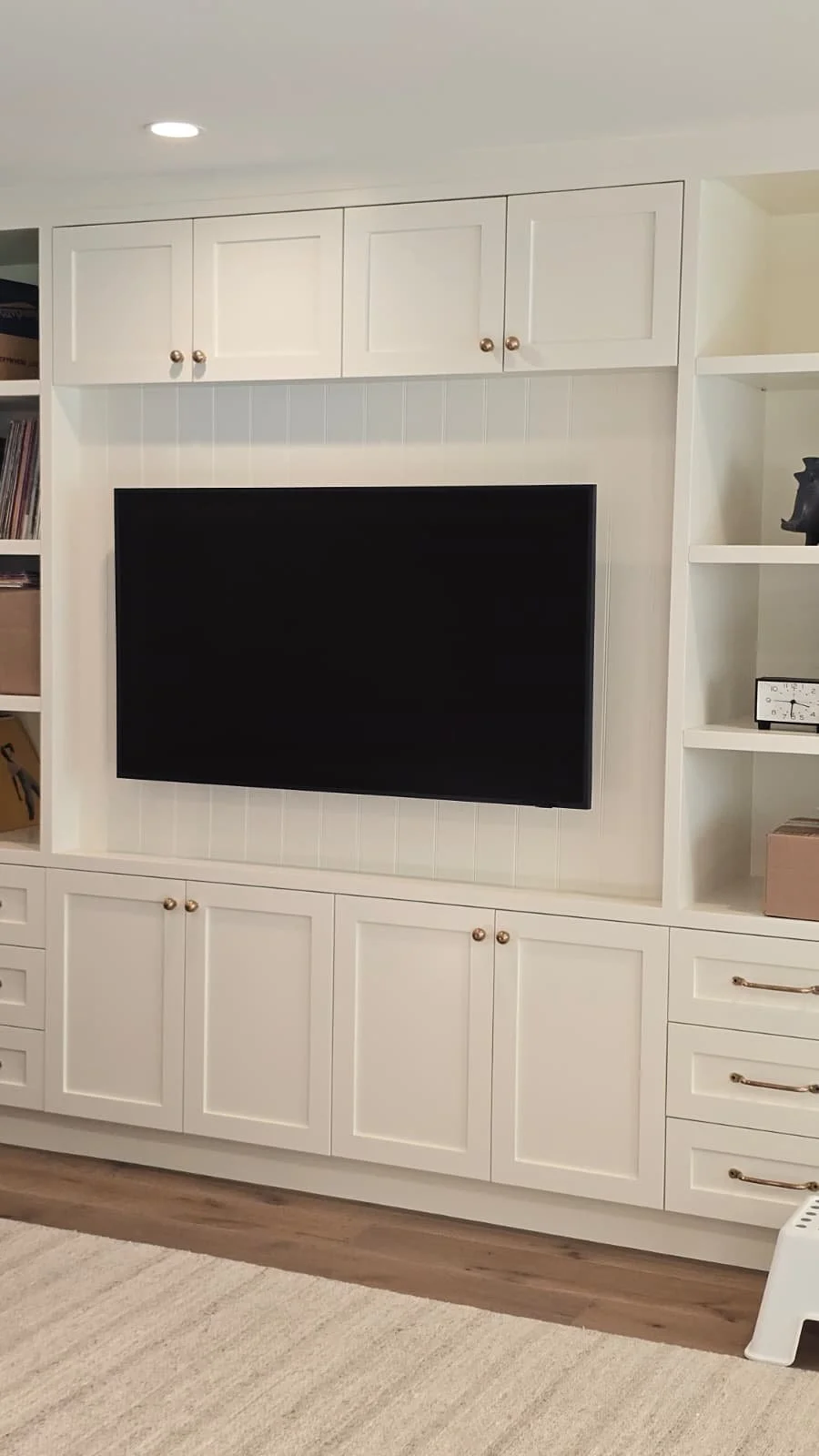 White built-in entertainment center with closed cabinets above and below, and open shelves on the sides. A large flat-screen TV is mounted in the center, with a beige rug on the light wood floor in front.