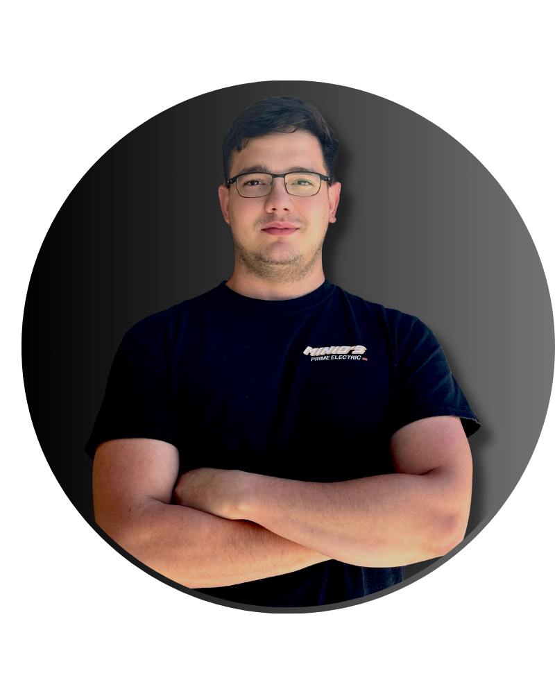 Young man with glasses and dark hair standing with arms crossed, wearing a black t-shirt that says 'NINNOS PRIME ELECTRIC'; dark background with a circular vignette effect.