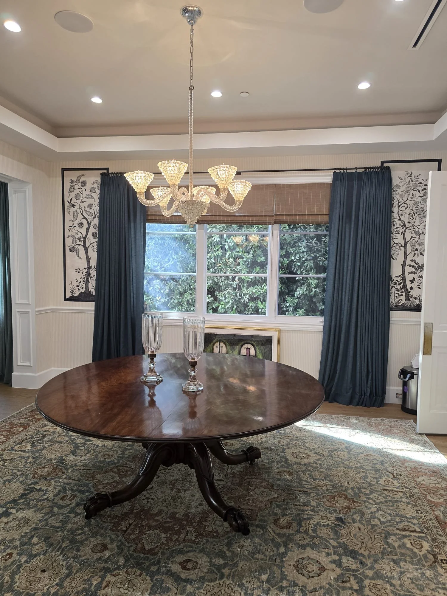 A dining room with a wooden round table, three tall glass candle holders, a large ornate chandelier, windows with blue curtains, wallpaper with a tree branch and leaf design, and a patterned rug.