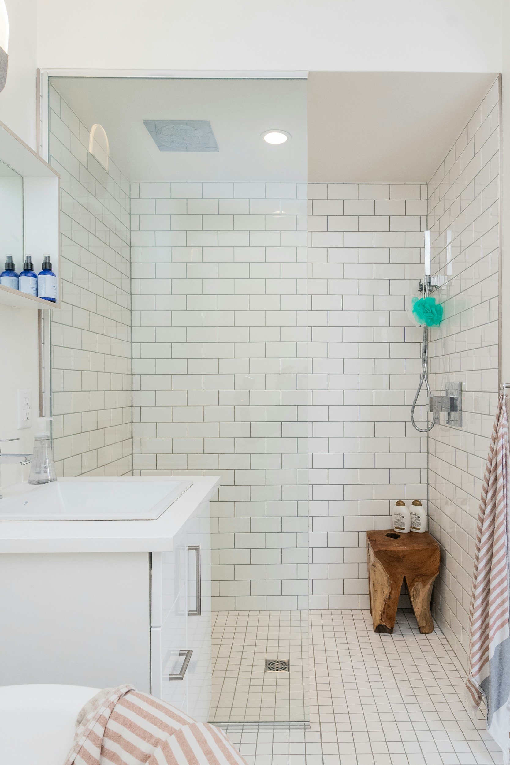 Modern bathroom with white subway tiles, a glass shower door, a wooden stool, and a sink with toiletry bottles on the counter.