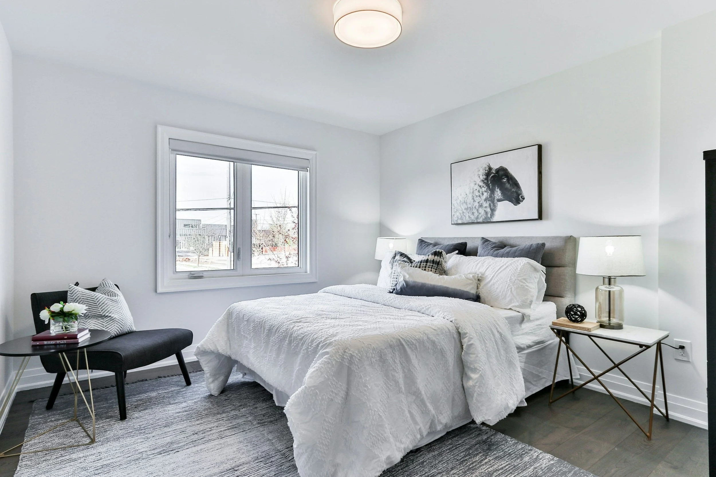 Modern bedroom with a neatly made bed, white bedding, and decorative pillows. A black and white sheep print hangs above the bed. A side table with a lamp and decor, a chair with a cushion, and a window are also visible.