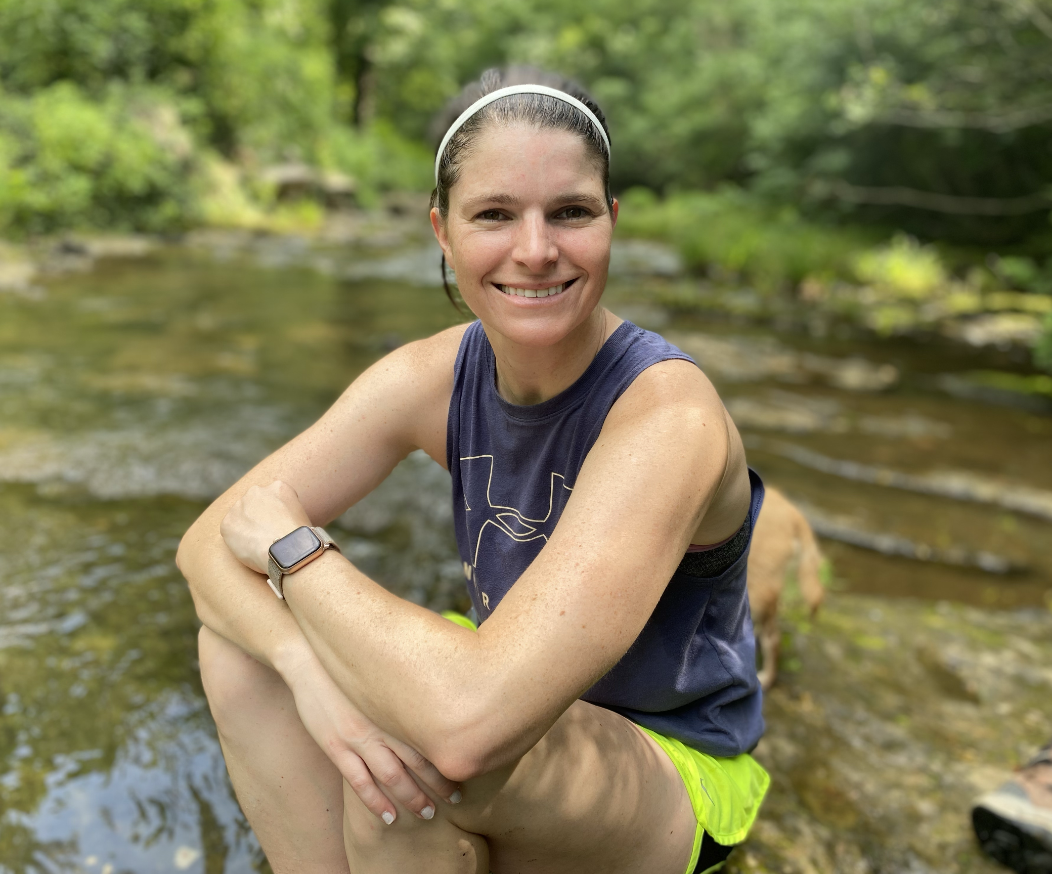 Women in athletic clothing smiling at camera while sitting by a river in a wooded area. Aimee Prudhomme. Iza Clara Healing testimonial. 