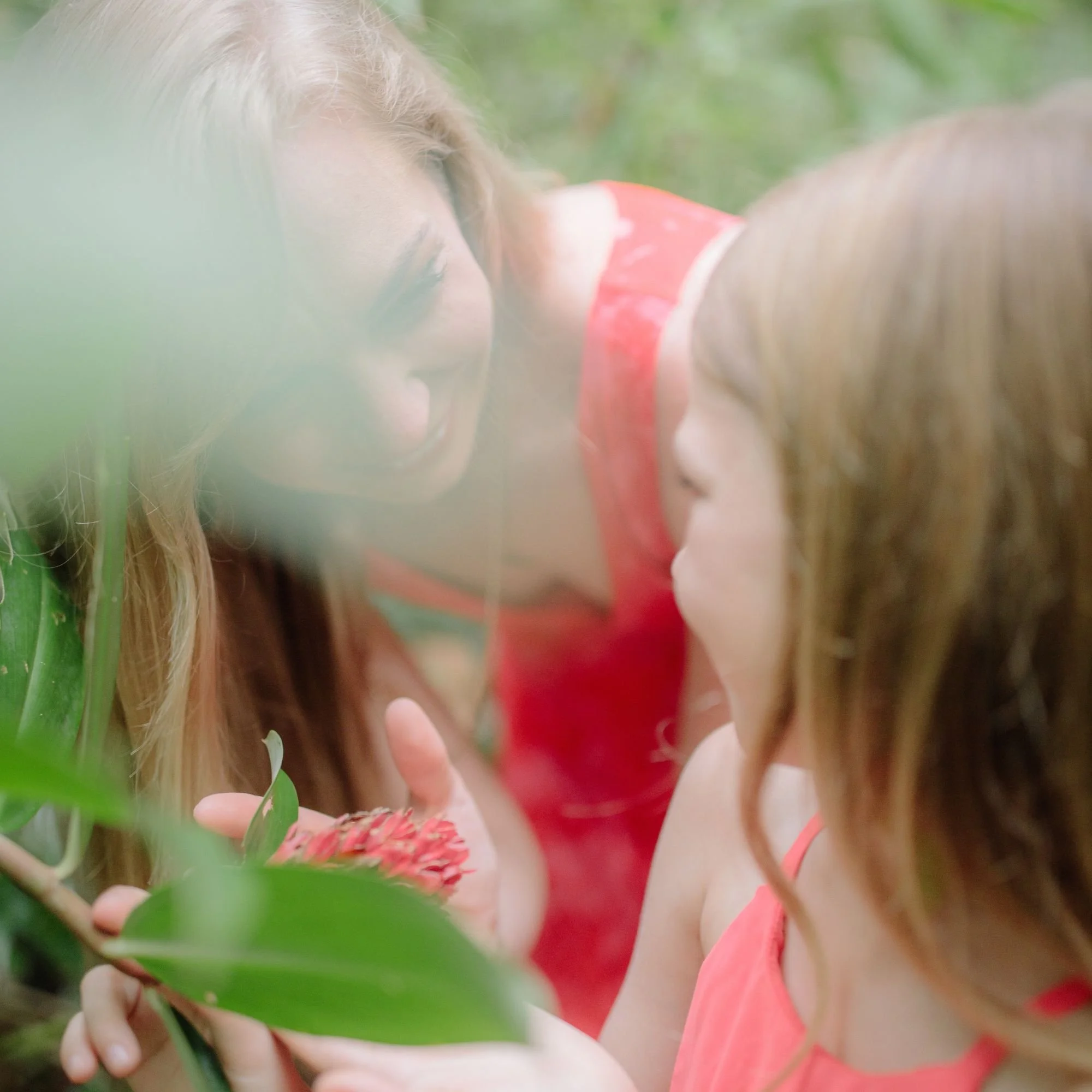 A woman and a child, both in red, smile at a red flower in a lush, green setting. The image conveys joy and connection with nature.