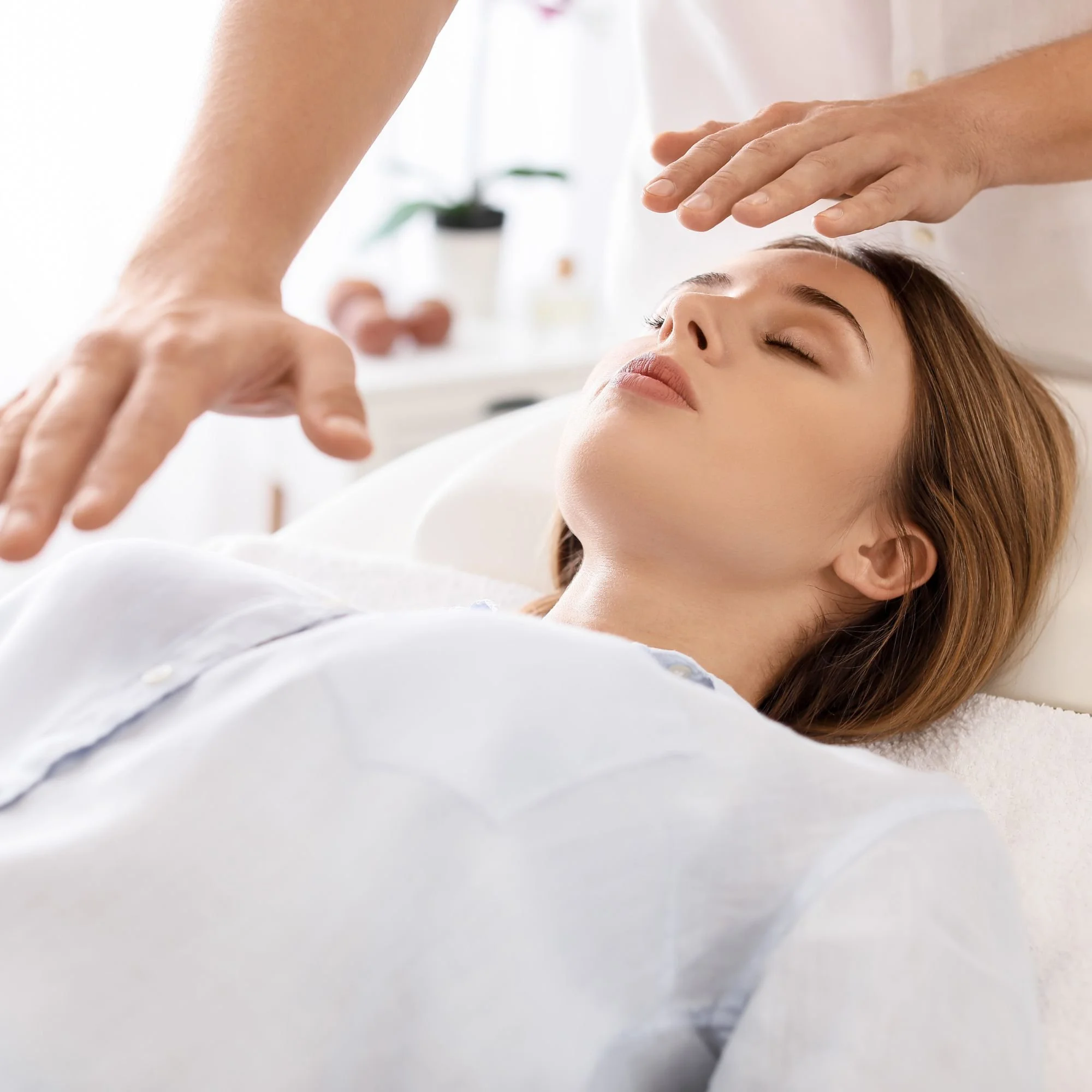A woman lies relaxed with eyes closed during a Reiki healing session. A practitioner's hands hover above her, creating a peaceful, calming atmosphere.
