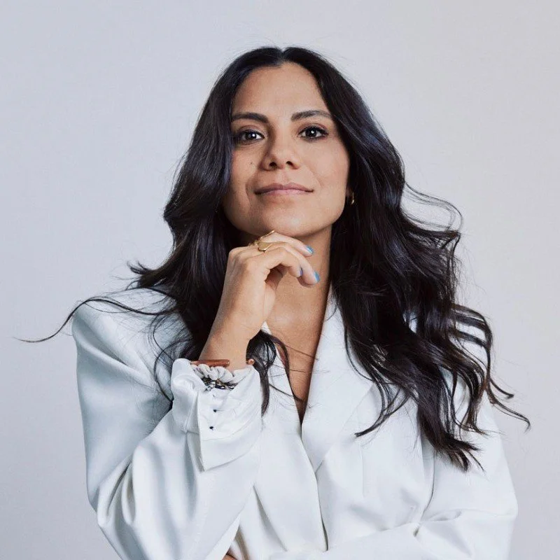 A woman with long dark wavy hair wearing a white blazer, looking confidently at the camera with her chin resting on her hand. Ana Maria Moreno, Founder of The Copal. Iza Clara Healing testimonial. 