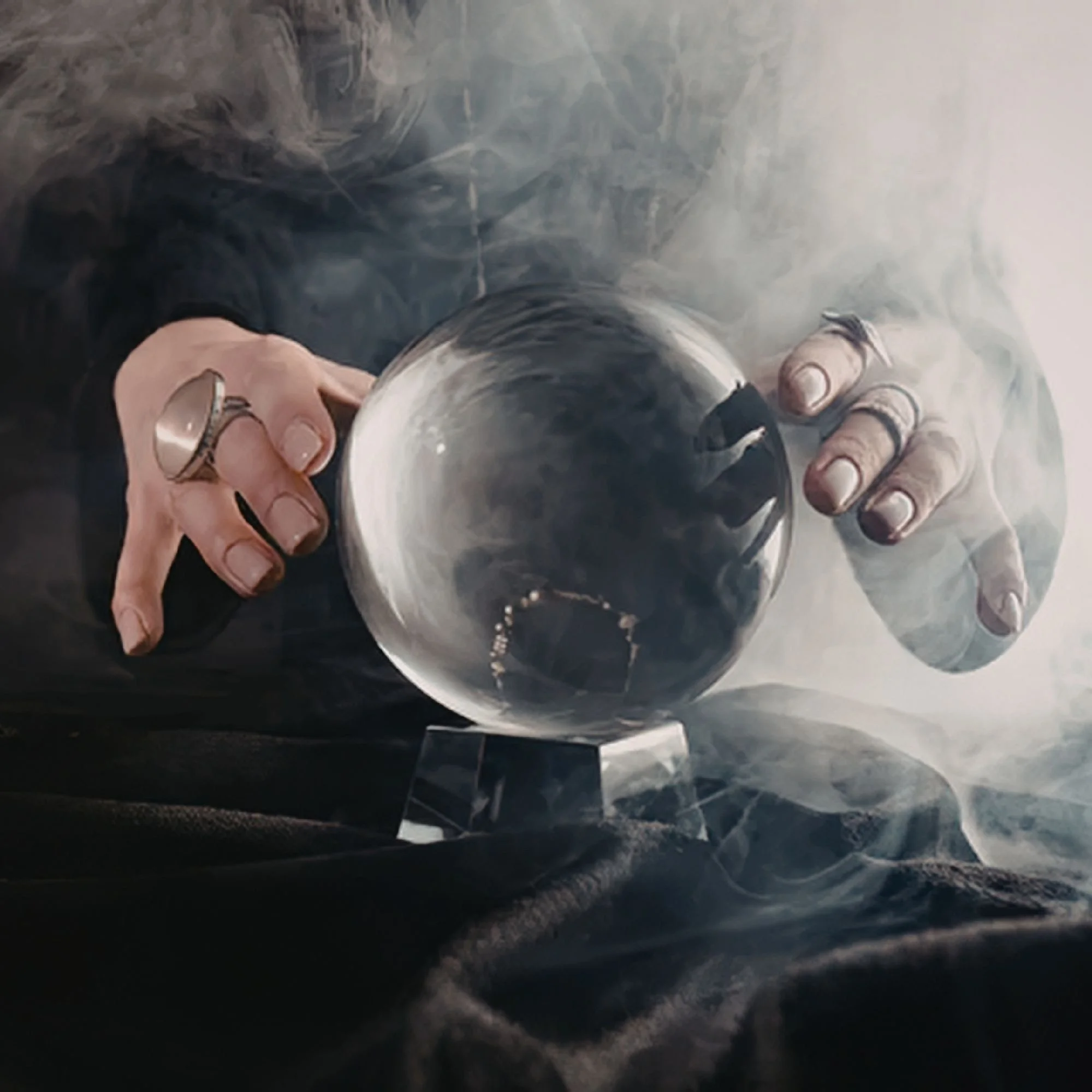 Hands adorned with rings hover over a smoky crystal ball on a dark cloth, creating a mysterious and mystical atmosphere.