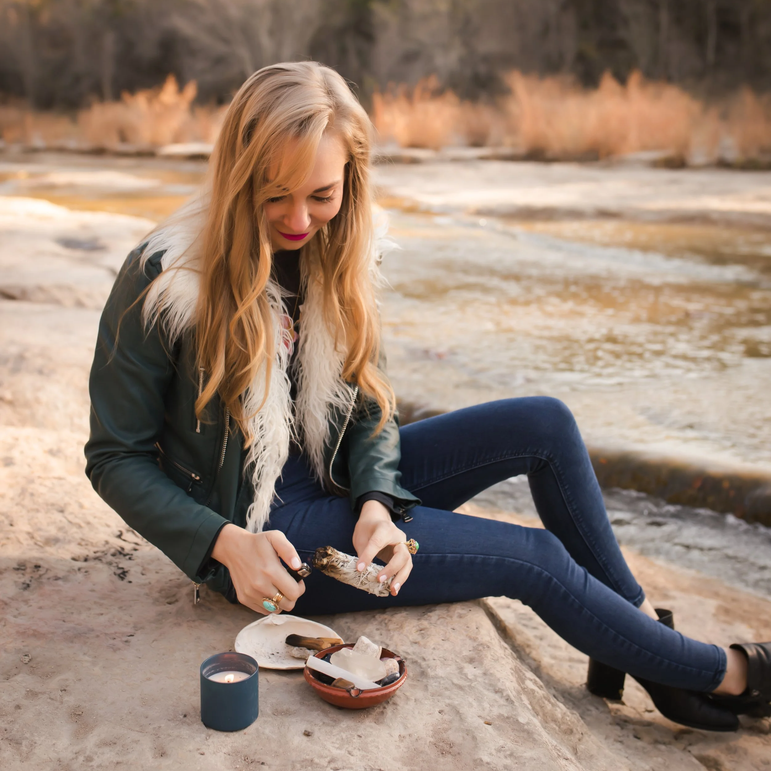 A woman with long blonde hair, wearing a faux fur-trimmed jacket, sits on a rock near a river, holding a smudge stick. The setting is peaceful with a mountainous backdrop.