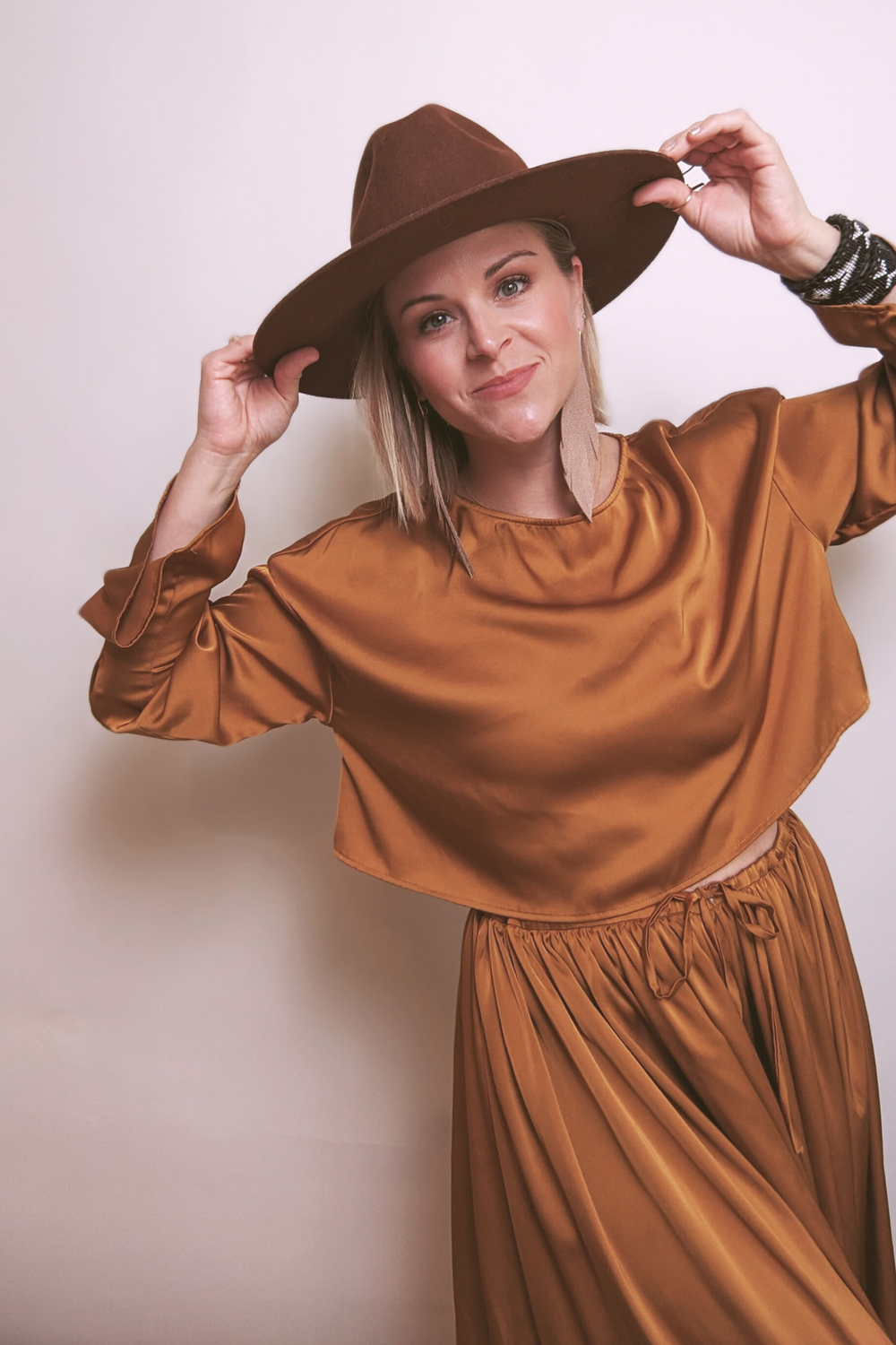 A woman with short blonde hair wears a brown wide-brimmed hat, a shiny brown satin top, and matching wide-leg pants. She is holding the brim of her hat with both hands and smiling at the camera against a plain white background. Kara Dockery 