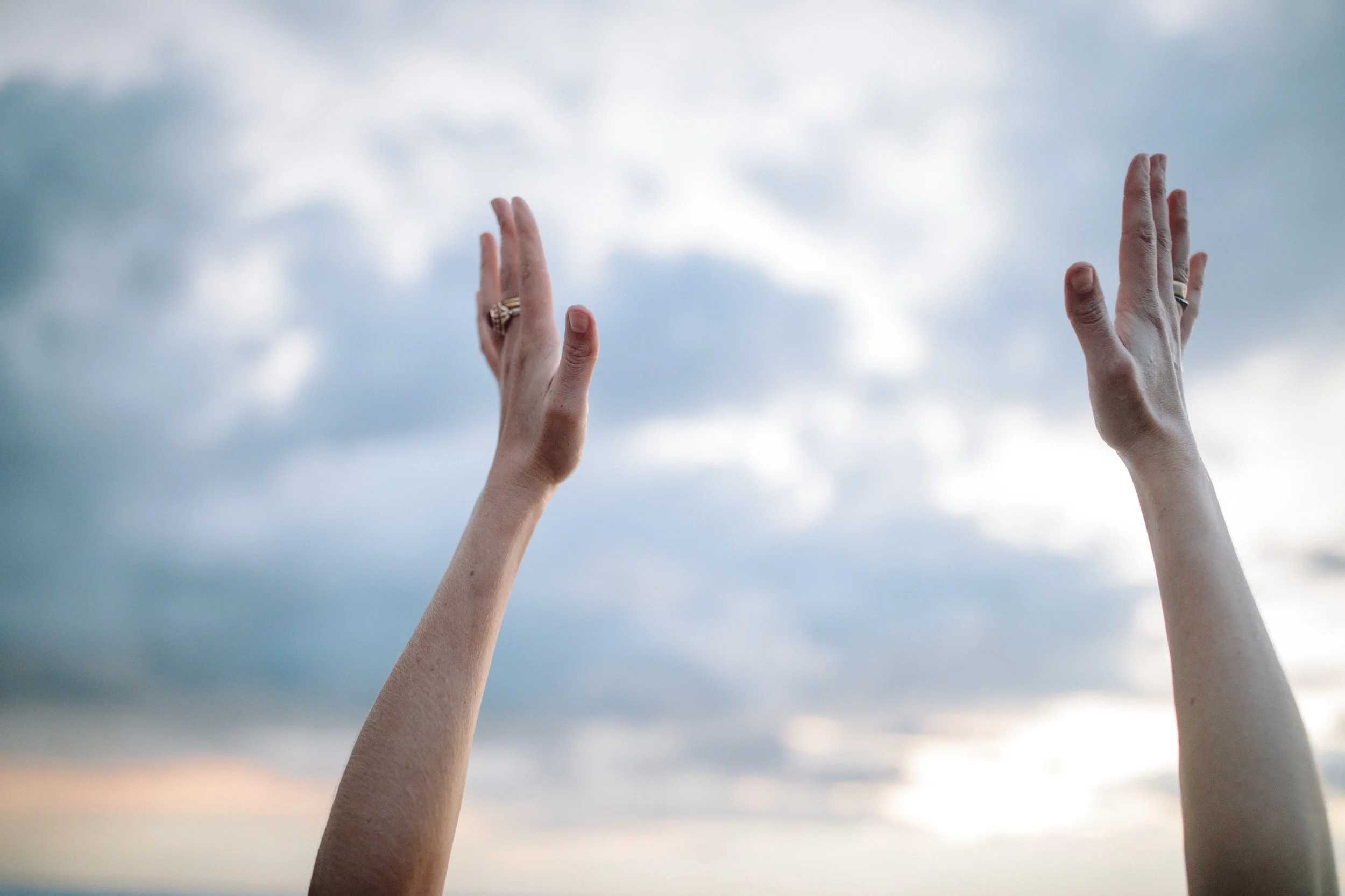 Two hands reaching up towards a cloudy sky during sunset. Iza Clara Healing Offerings. 