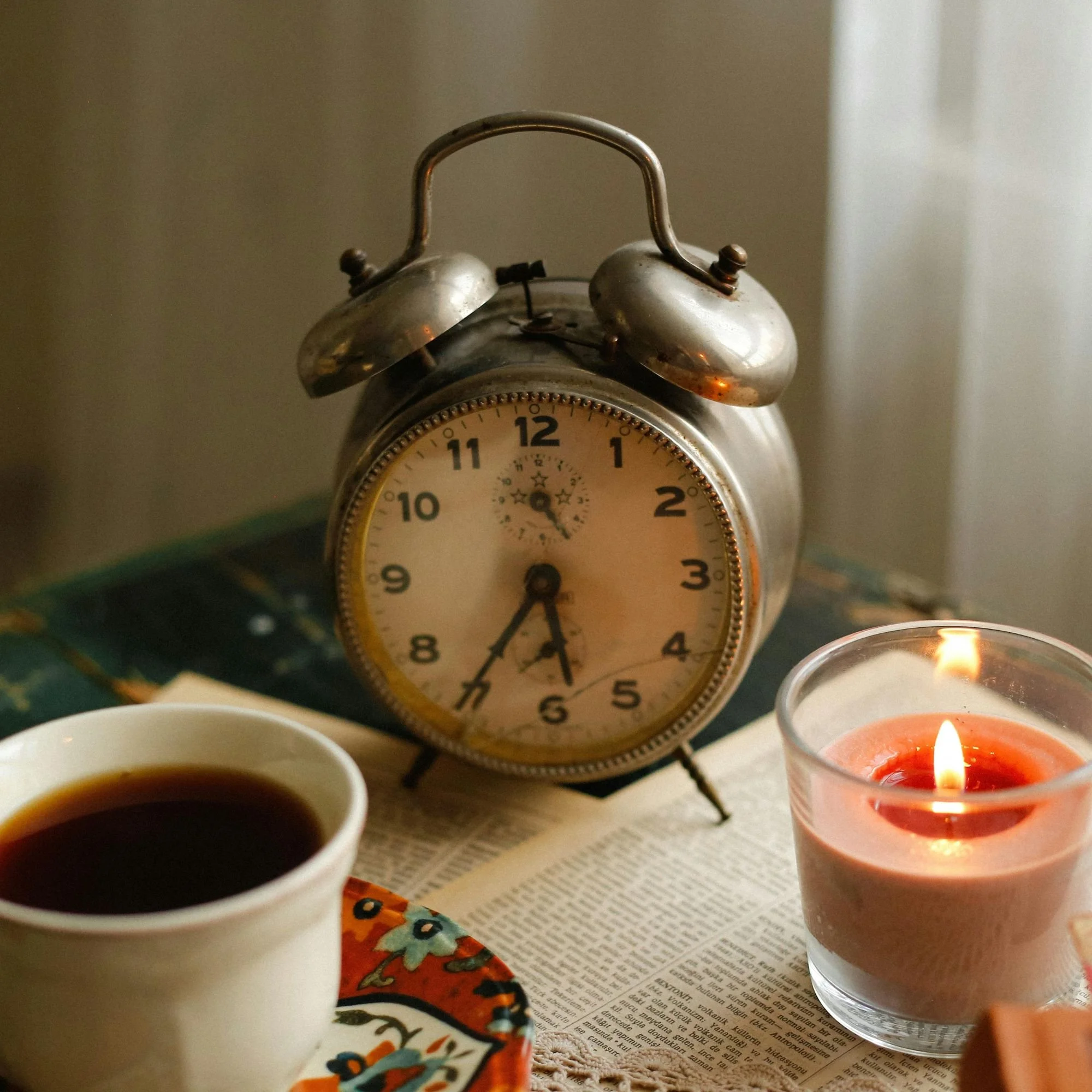 A vintage alarm clock shows 6:30 next to a lit candle and a cup of coffee on a table with a newspaper. The scene conveys a cozy, nostalgic morning.