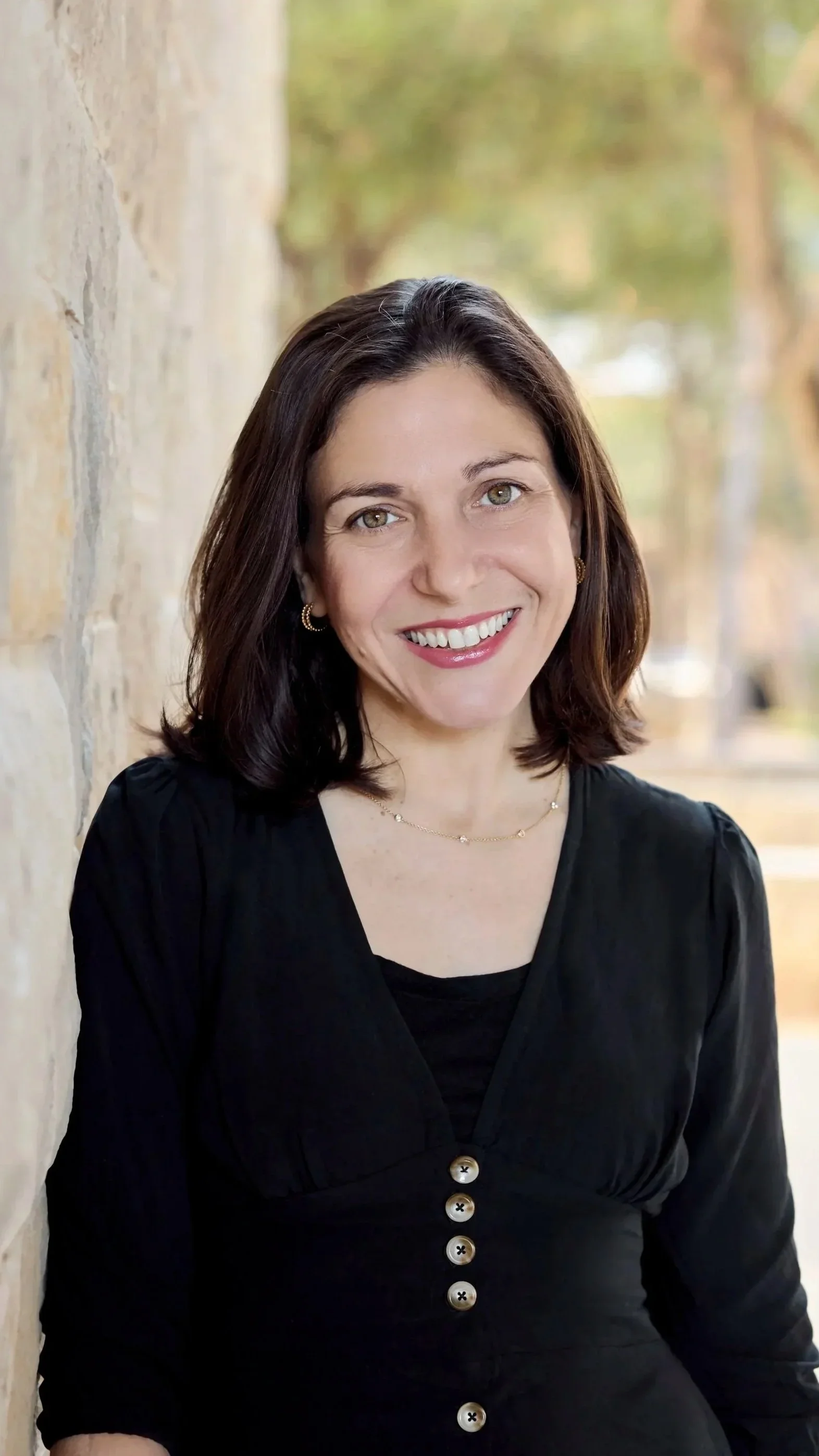 A woman with dark hair smiles warmly at the camera. She is wearing gold hoop earrings. The background is softly blurred, with a light stone wall on the left.