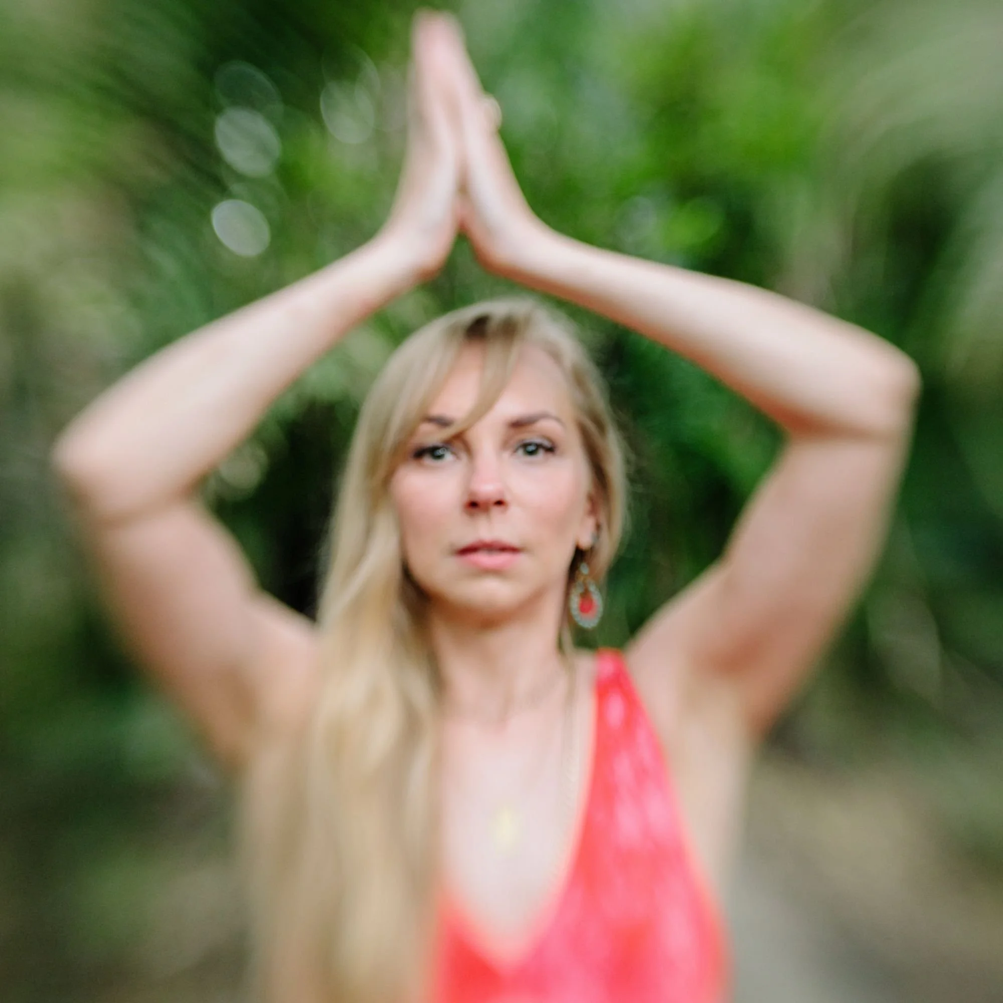 A woman with long blonde hair stands in a serene forest setting, wearing a red dress. Her hands are clasped above her head in a yoga pose, conveying calmness.