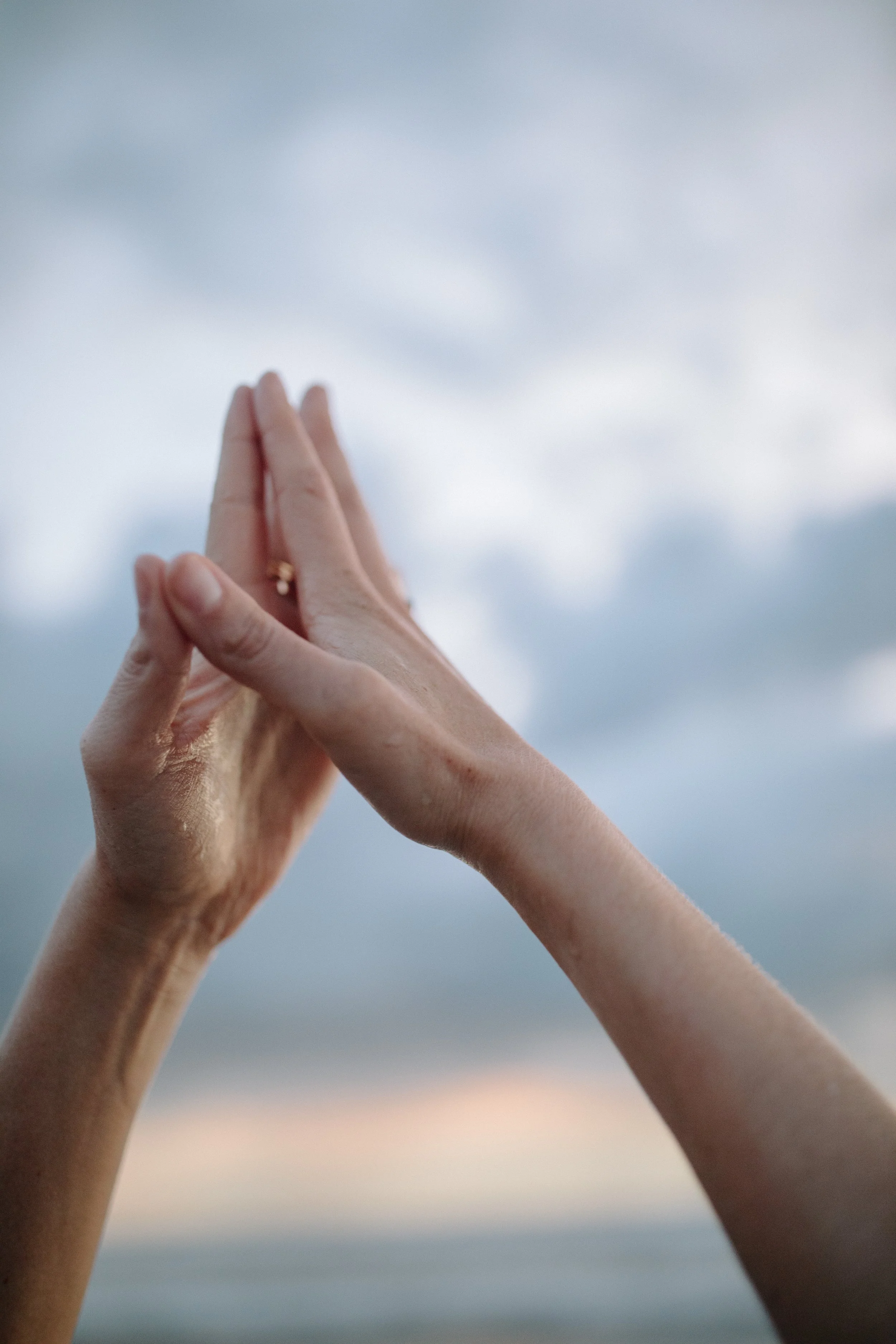 Two hands pressed together in a prayer or namaste gesture against a cloudy sky background. Iza Clara Healing Offerings. 