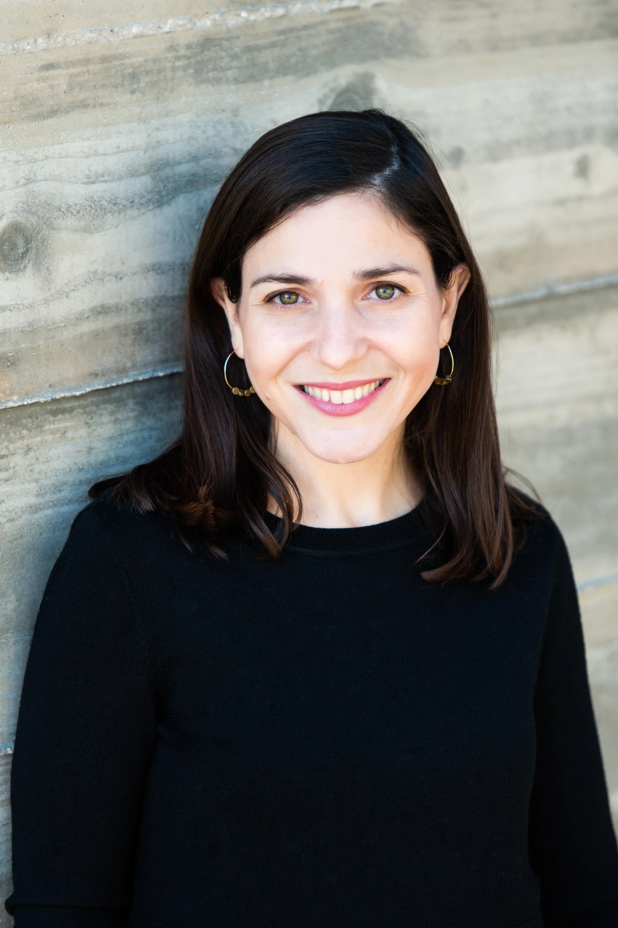 A woman with dark brown hair, green eyes, and hoop earrings smiling at the camera, standing against a wooden wall. Rachel Glaser, LCSW. Iza Clara Healing testimonial. 