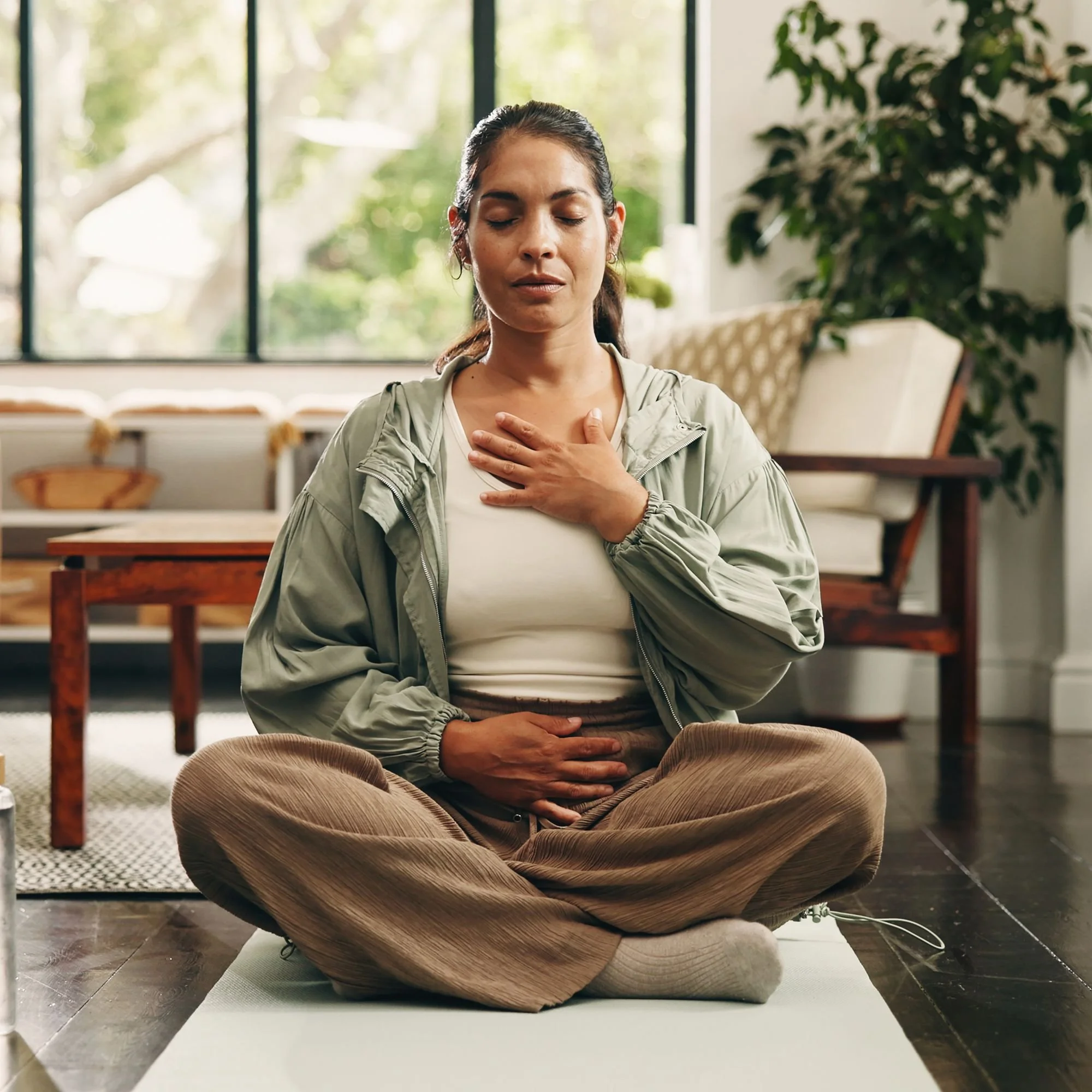 A woman sits cross-legged on a yoga mat in a cozy living room, eyes closed, one hand on her chest and the other on her belly, conveying calm and mindfulness.