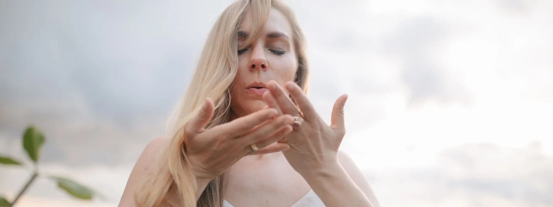 A woman in a white dress stands outdoors with eyes closed, gently blowing on her hands. The background is a serene beach setting, conveying calmness.