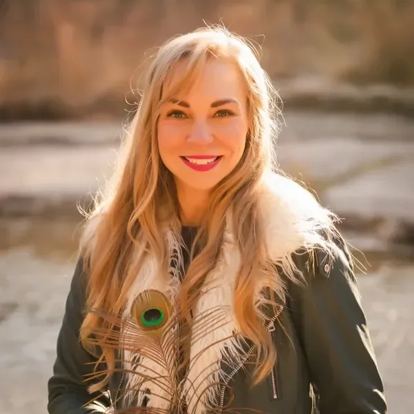 A woman with long blonde hair smiles warmly, holding a peacock feather. She's wearing a dark jacket with a white fur collar, set against a blurred outdoor background.
