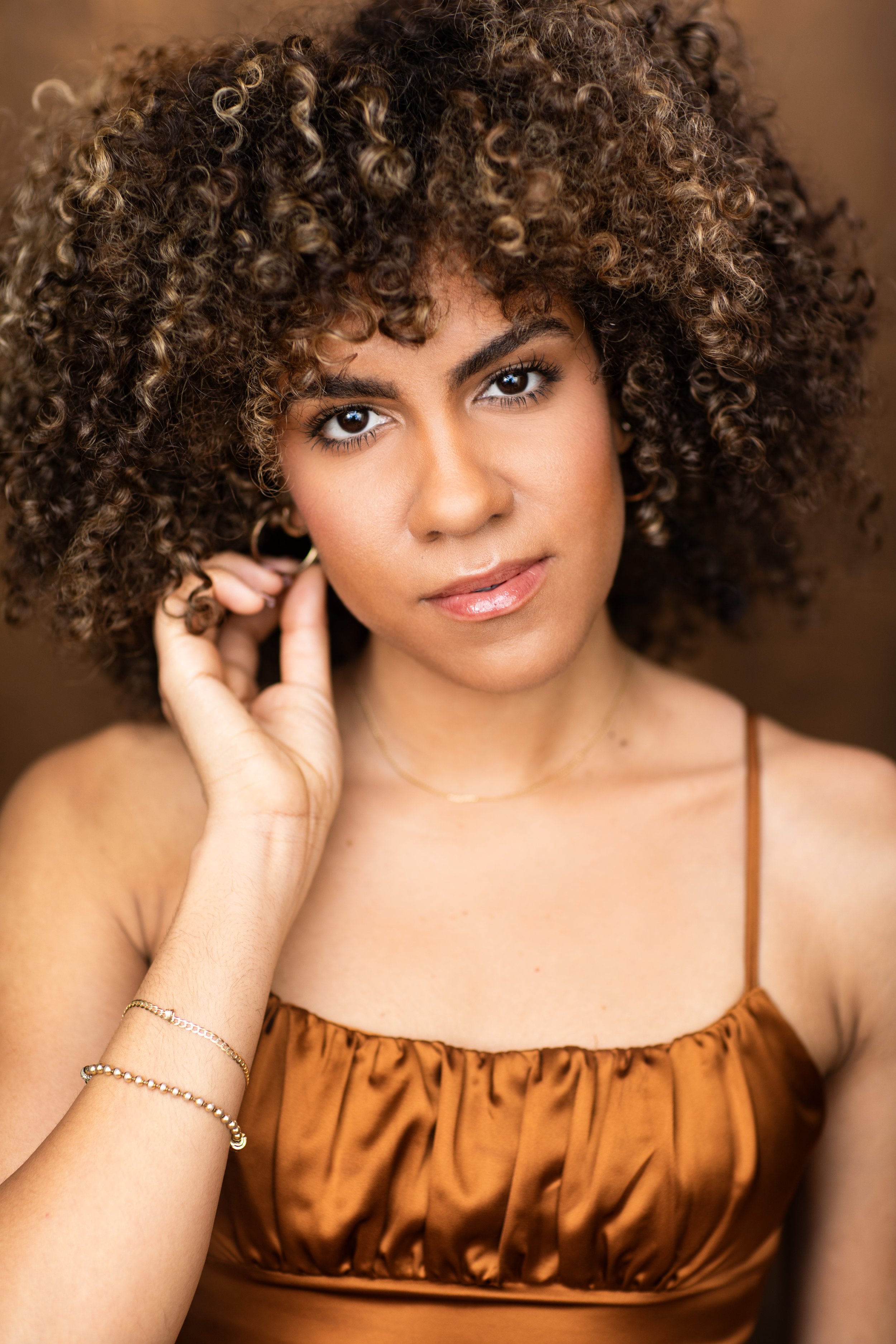 Jolimar wearing a bronze scrunched top, staring at the camera touching her right earing