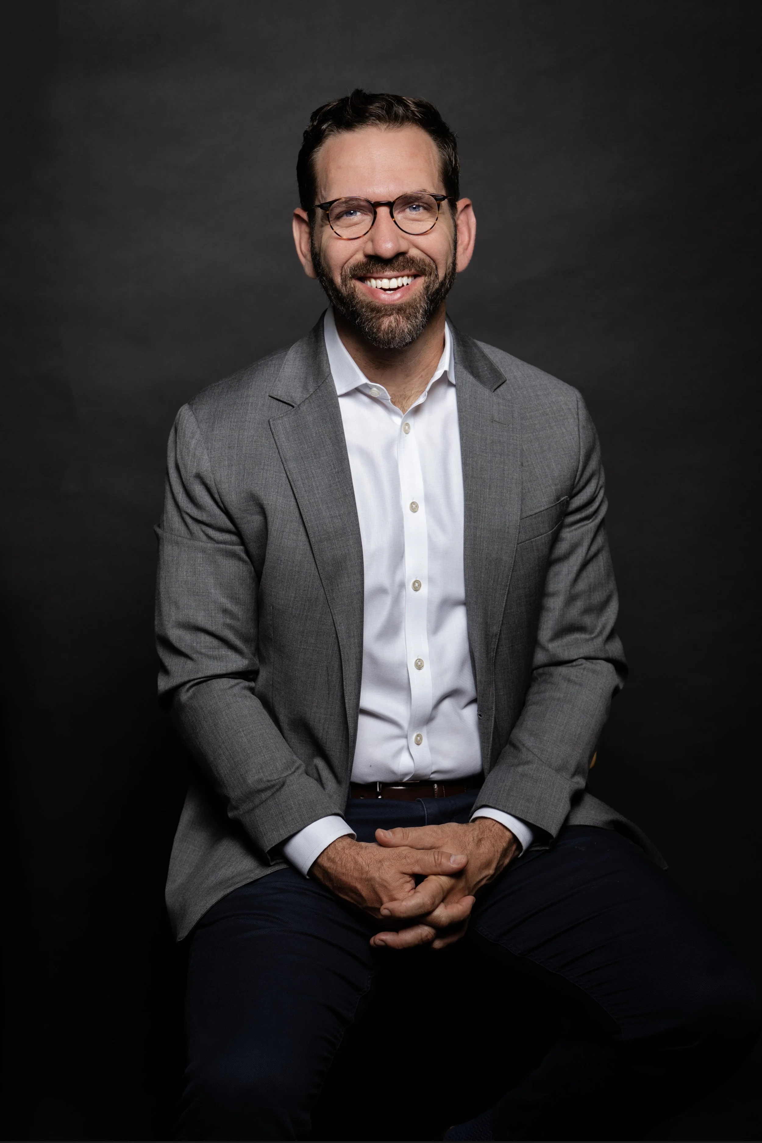 A man with glasses, a beard, and dark hair, wearing a gray suit and white shirt, smiling and sitting against a dark background for a corporate headshot done in a studio