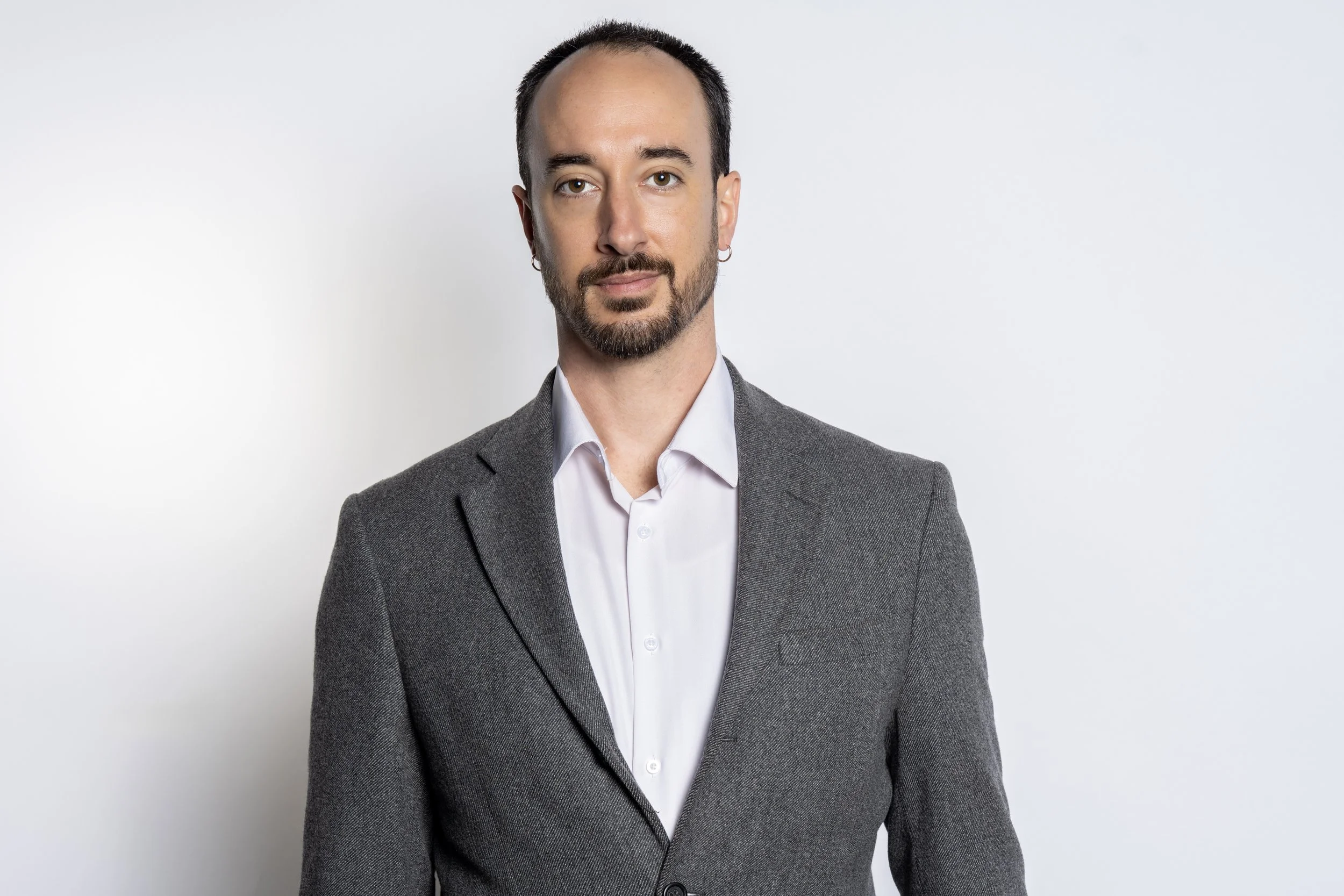 A man with a beard and earrings wearing a gray blazer and white shirt, standing against a plain light background for a corporate headshot done in a studio