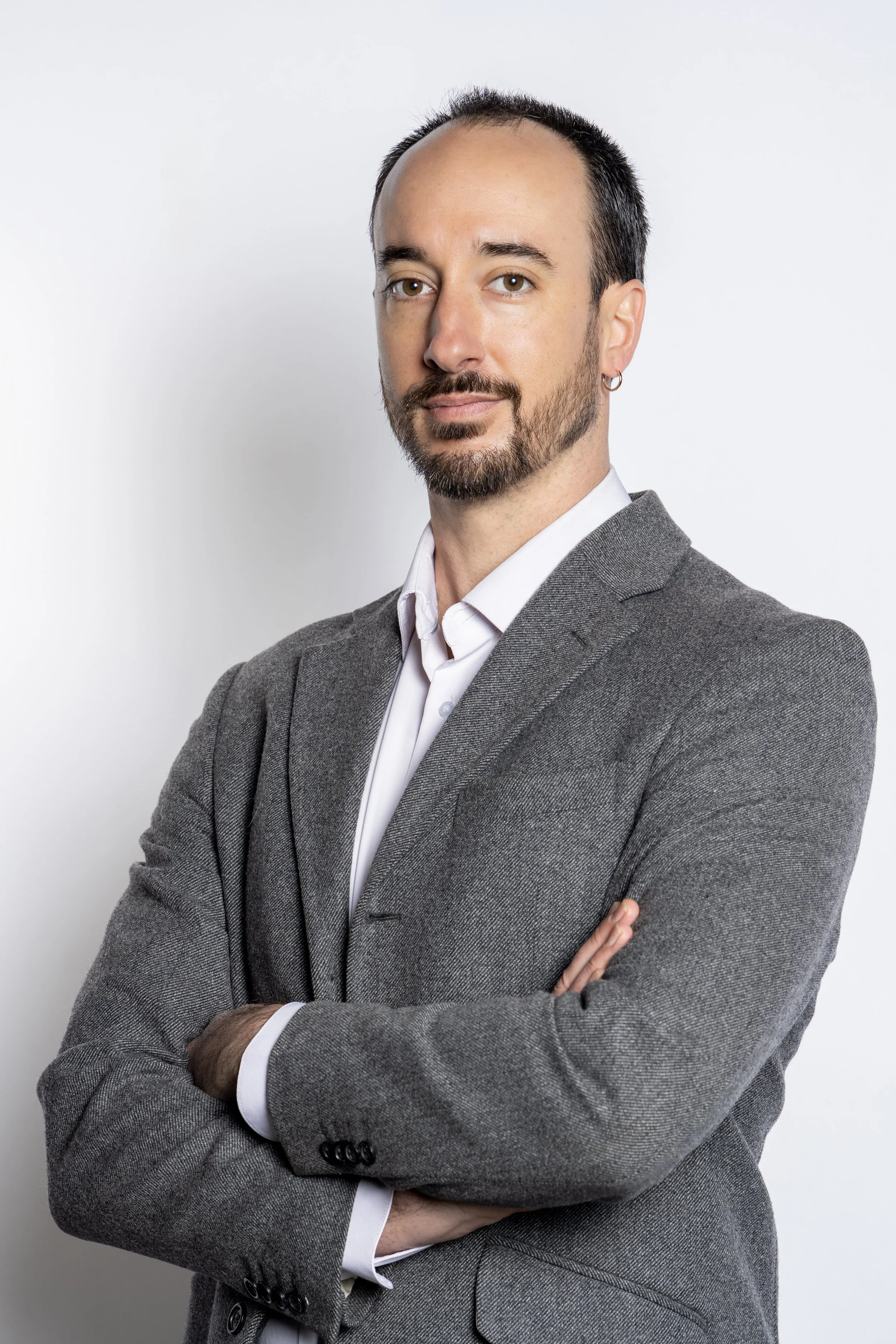 A man with light skin, a beard, and a hoop earring in his left ear, wearing a gray suit and white shirt, standing against a plain white wall with arms crossed for a corporate headshot done in a studio